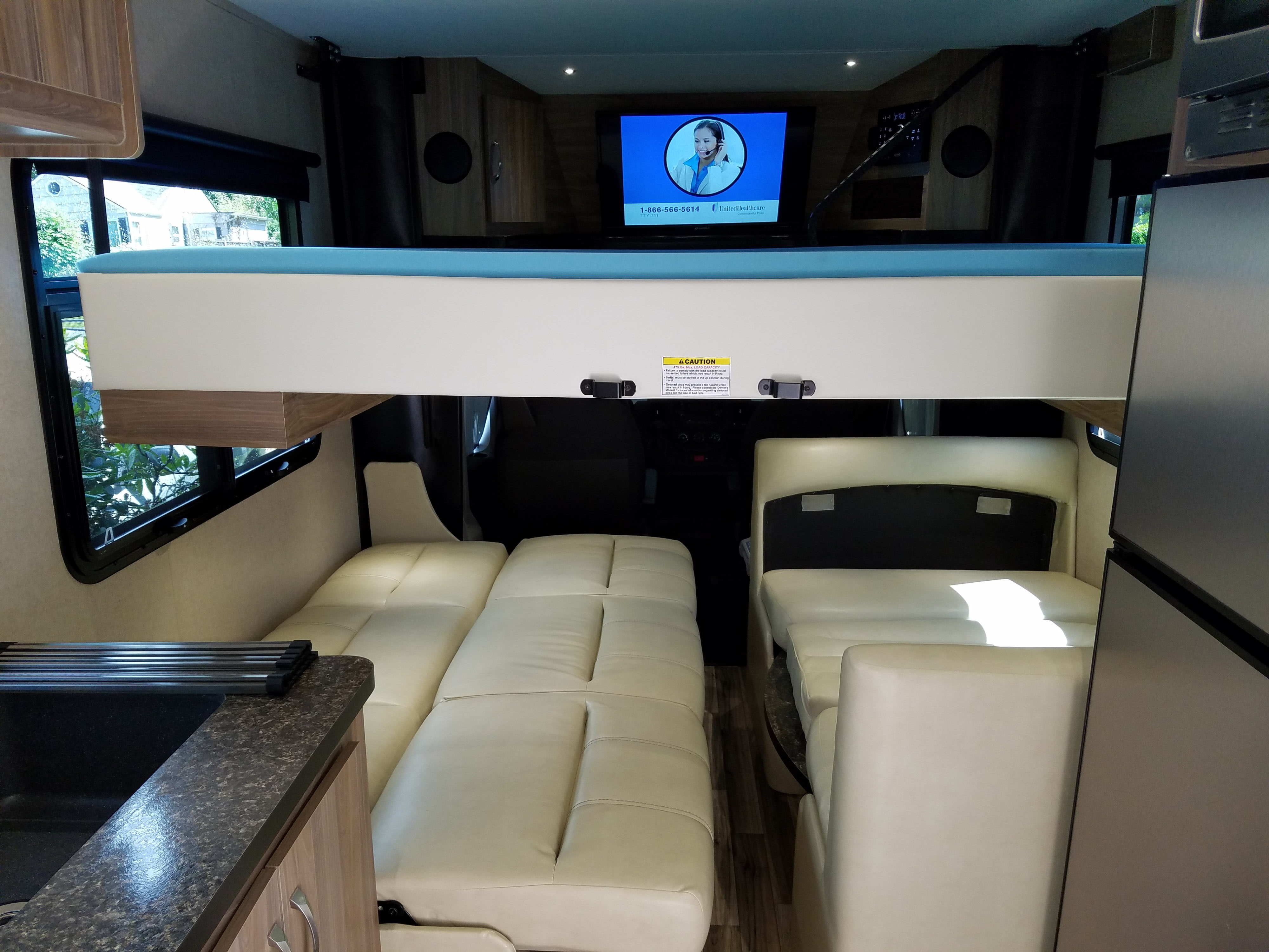 RV rental interior with an elevated bunk bed, convertible lounge seating, compact kitchen area with sink, and fridge.. Dynamax REV 24RB 2018