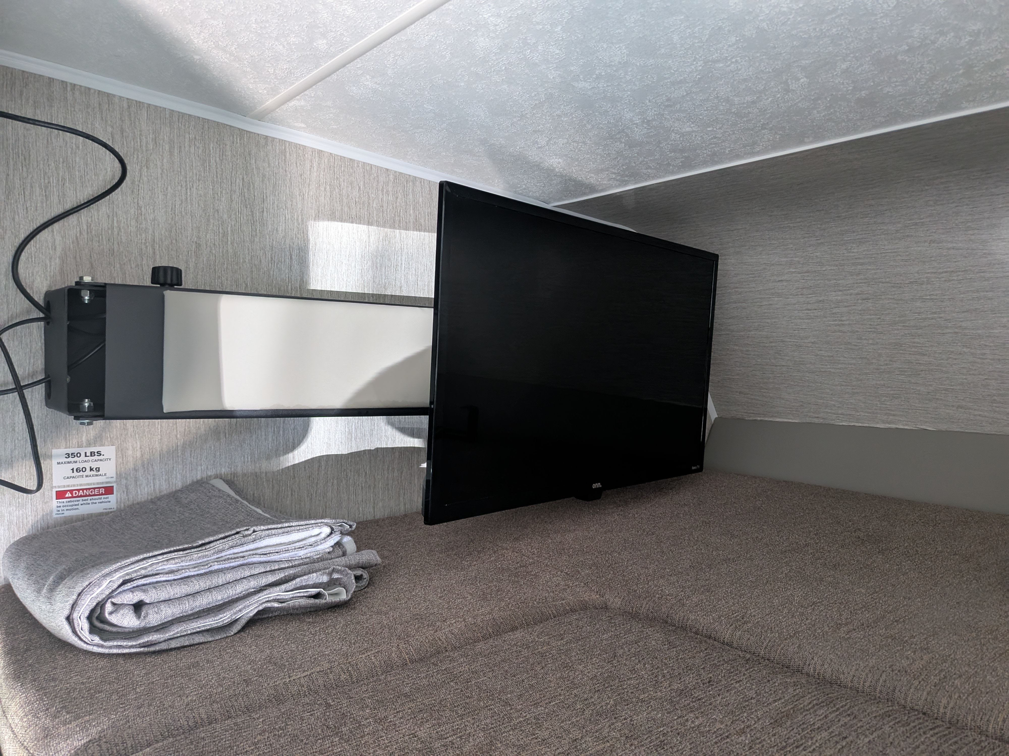 RV rental interior features: a mounted TV above a bed with a folded blanket and a 350lb capacity warning label.. Coachmen Freelander 2021