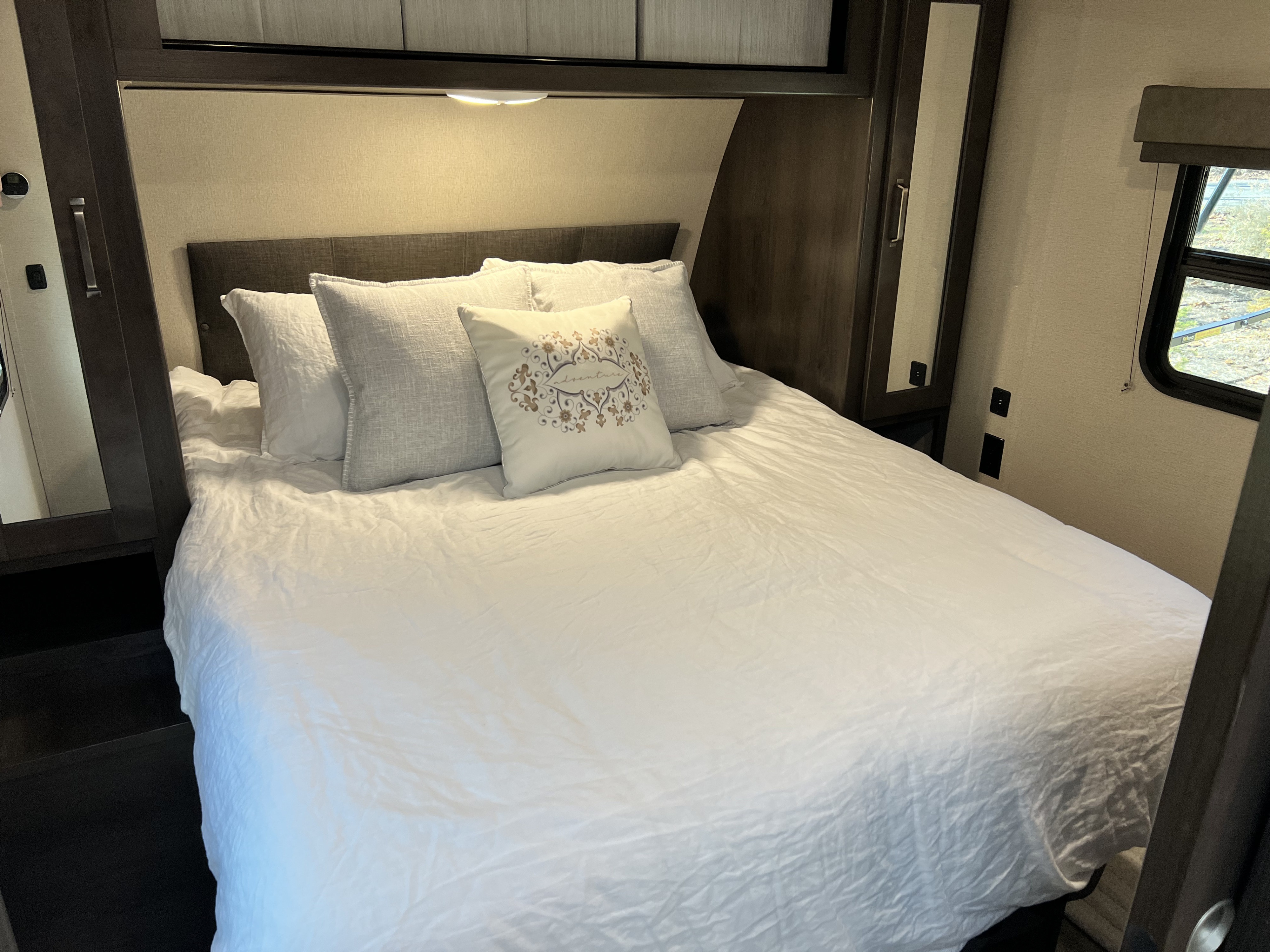 Cozy RV rental bedroom featuring a white bed with pillows, dark built-in storage, and a window, ideal for travel.. Grand Design Transcend Xplor 2022