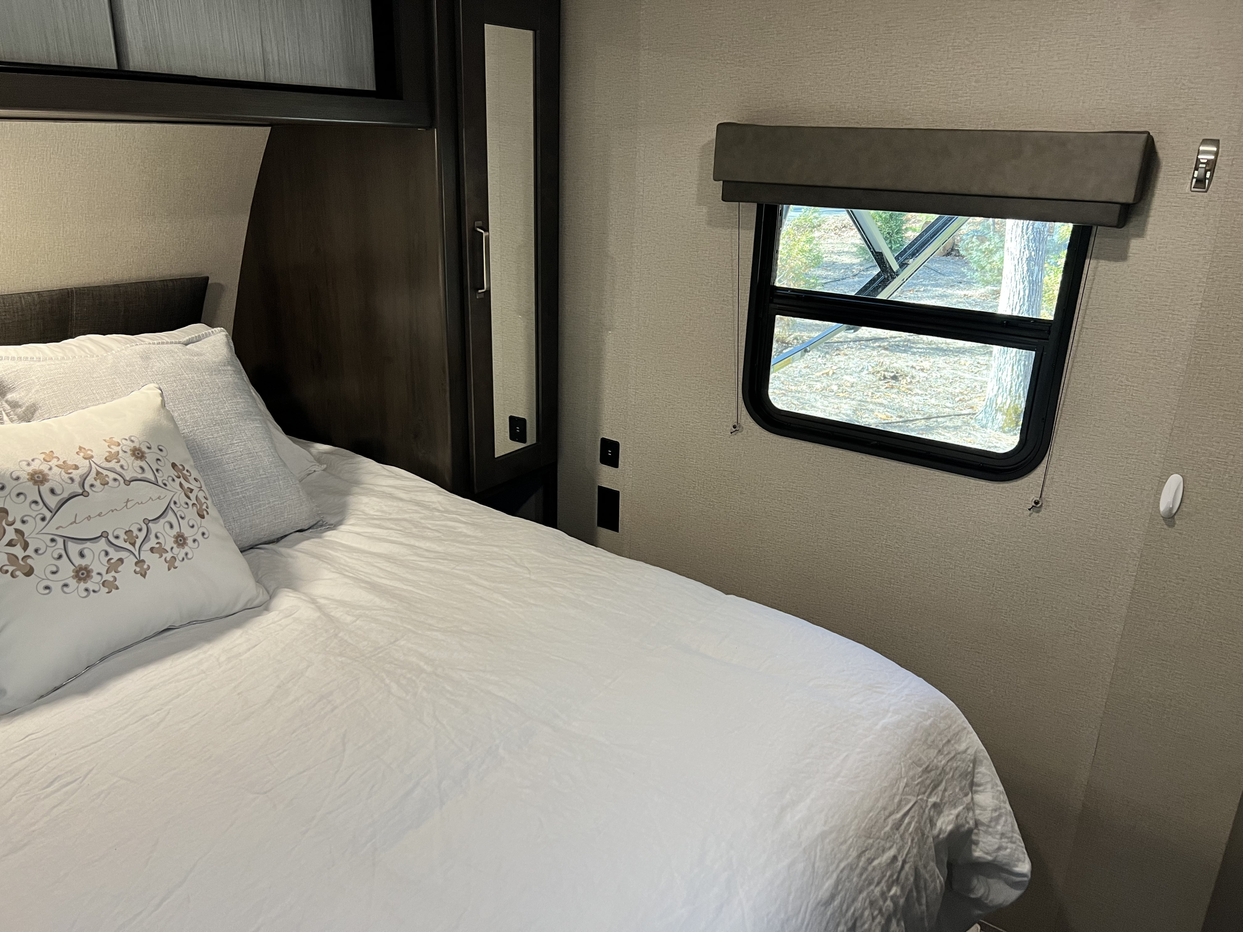 Cozy RV rental bedroom featuring a comfortable bed with white linens, decorative pillows, and a window with an outdoor view.. Grand Design Transcend Xplor 2022