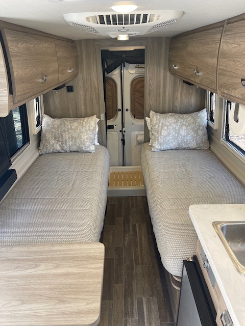 RV rental interior view: two twin beds with white textured comforters, light wood cabinets, and compact kitchen counter with sink.. Winnebago Travato 2023