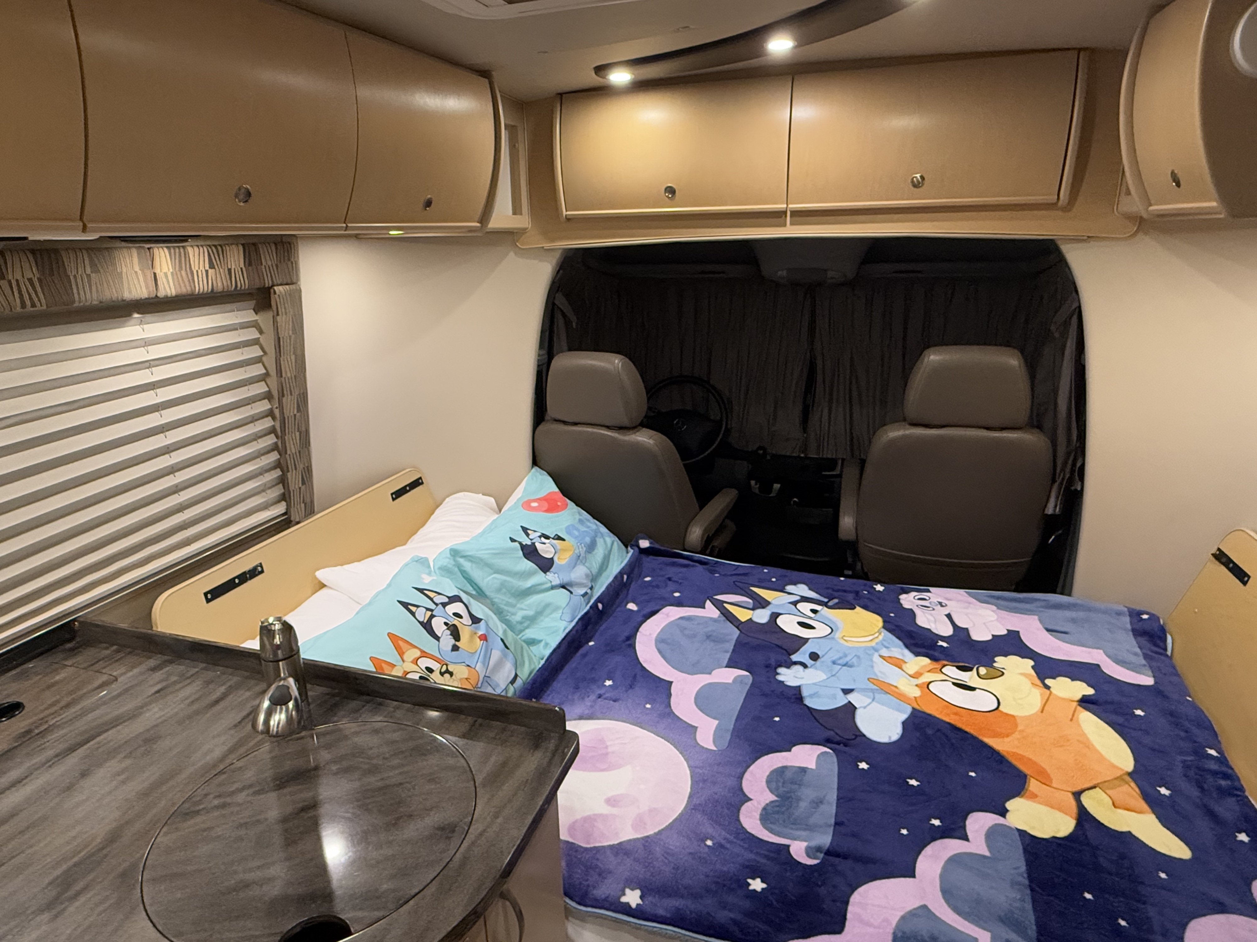 RV rental's cozy interior: Bluey-themed bed, small kitchen area, and overhead storage. Driver's cabin visible in front.. Leisure Travel Vans Serenity 2013