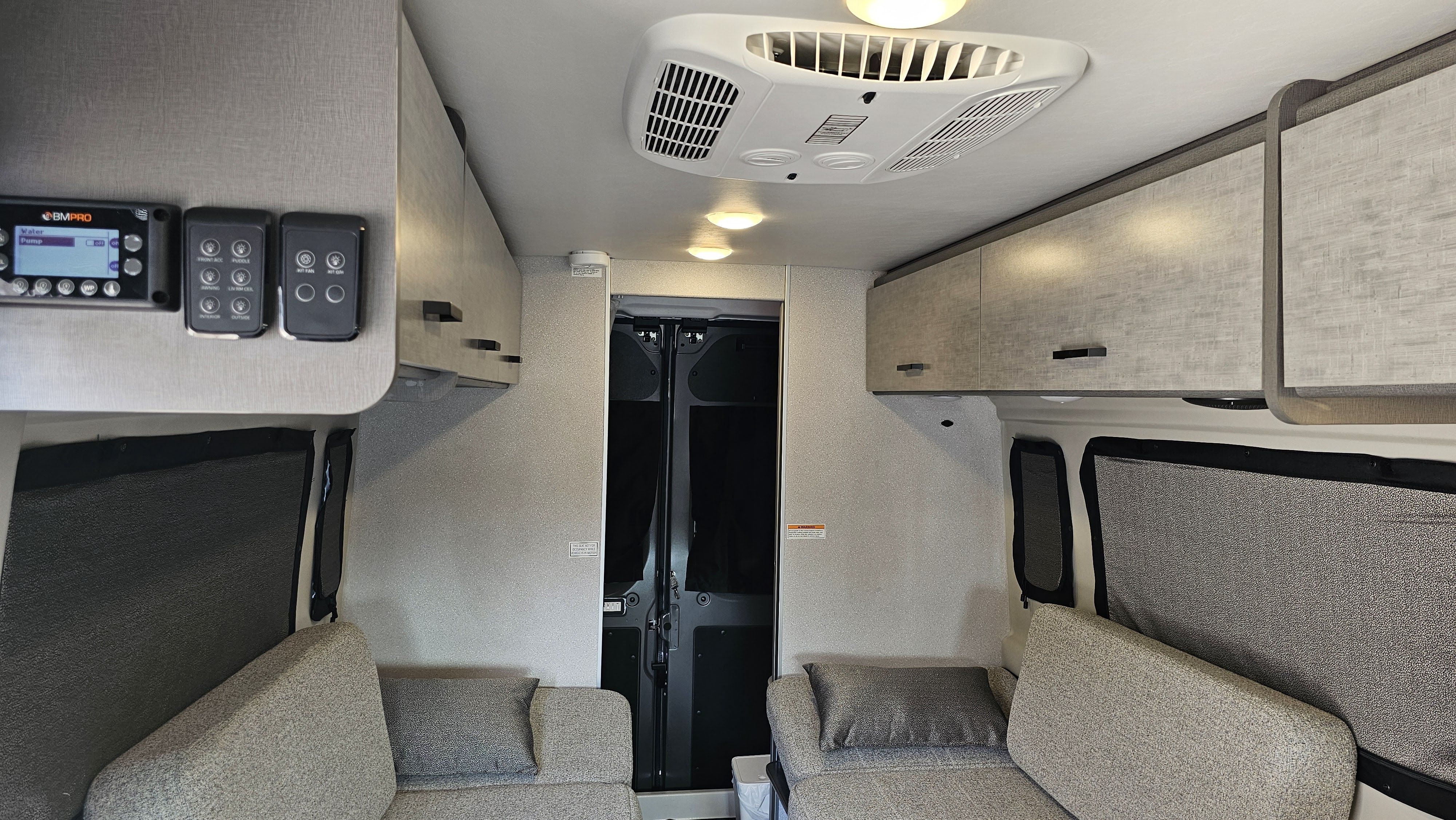 A bright RV rental interior showcases comfy seating with pillows, overhead cabinets, ceiling AC, and a control panel.. Thor Motor Coach Dazzle 2025