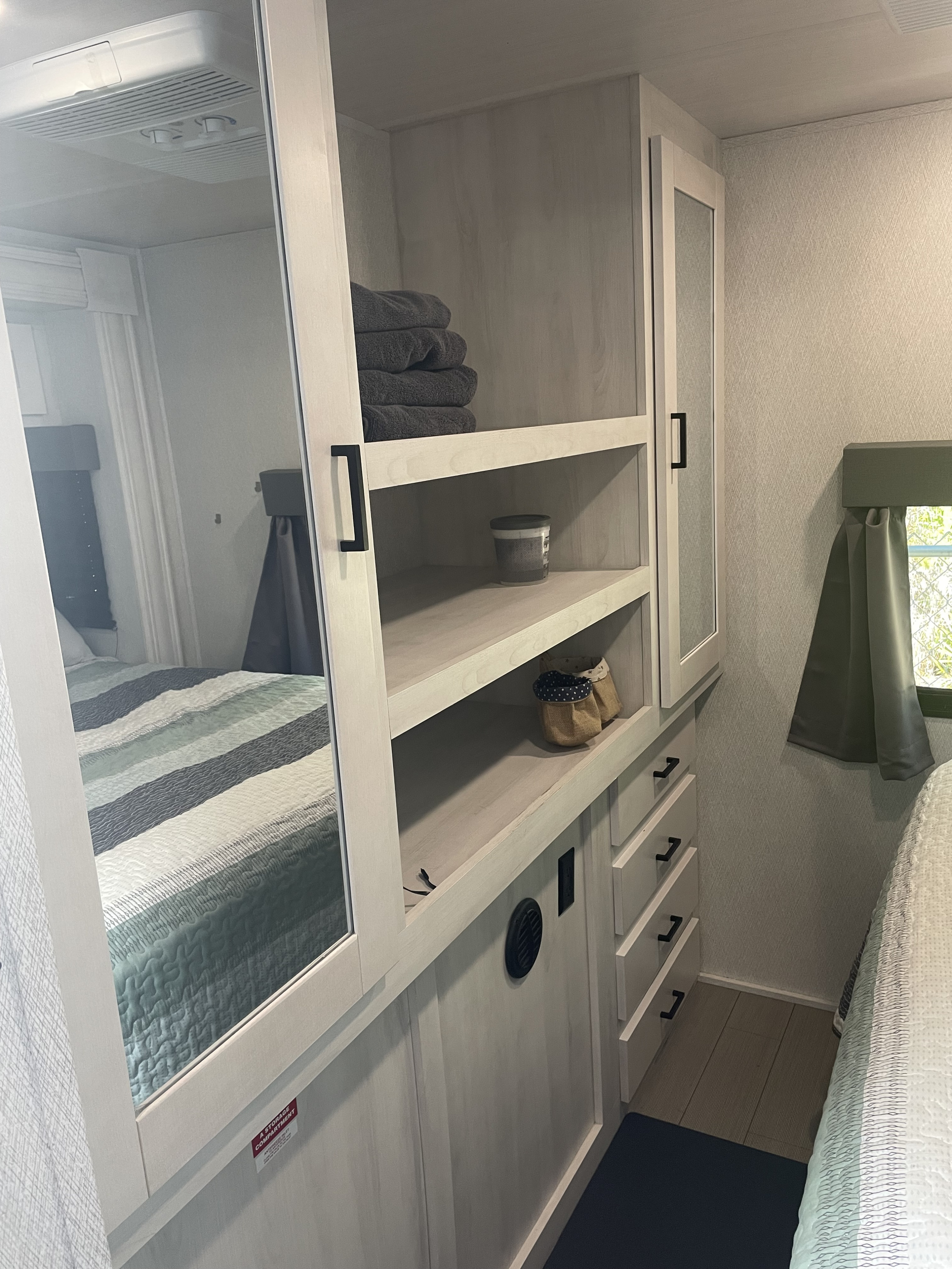 Inside an RV rental: light wood storage with open shelves, towels, drawers, and a mirrored closet reflecting the bed.. East to West Entrada 2023
