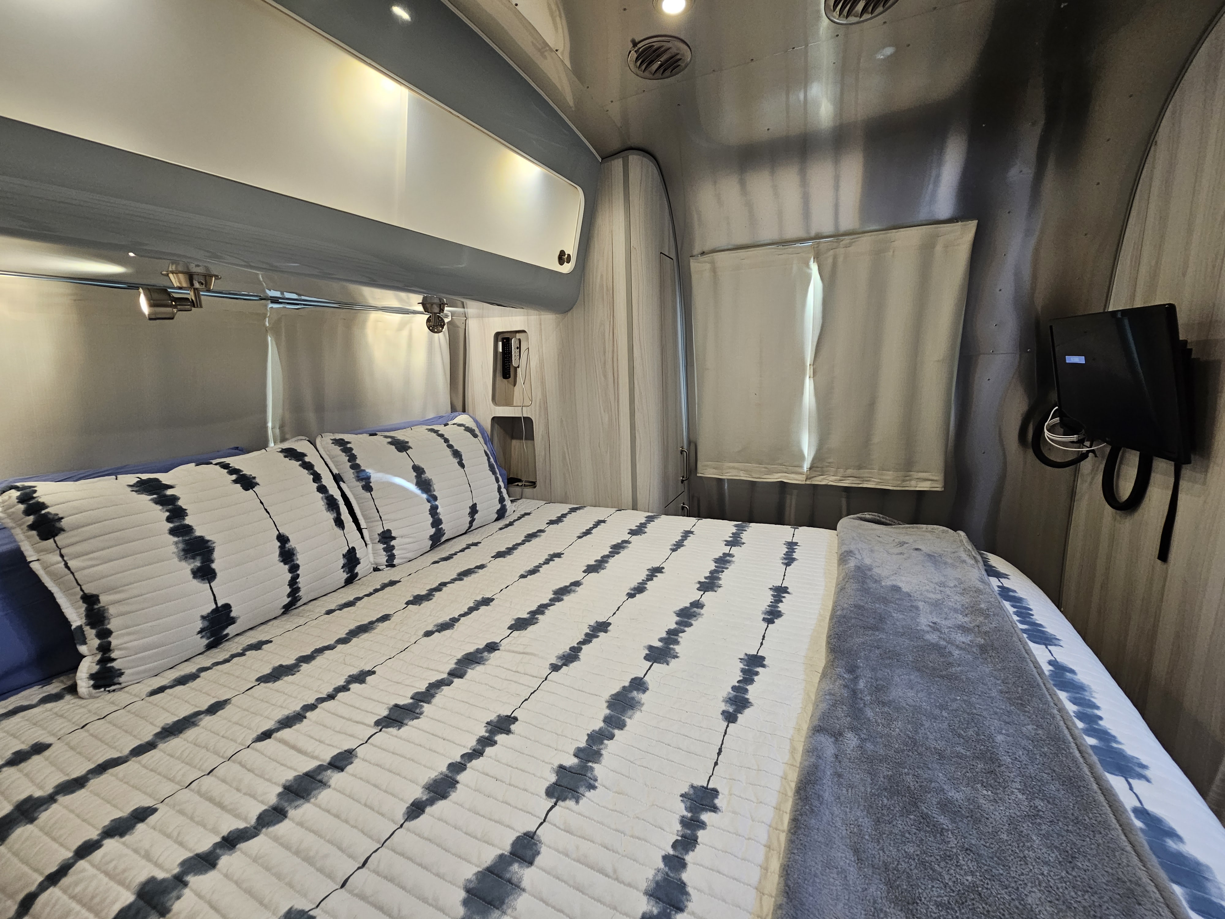 Inside an rv rental, a cozy bedroom features a bed with white and blue tie-dye bedding, gray blanket, and sleek silver walls.. Airstream International 2022