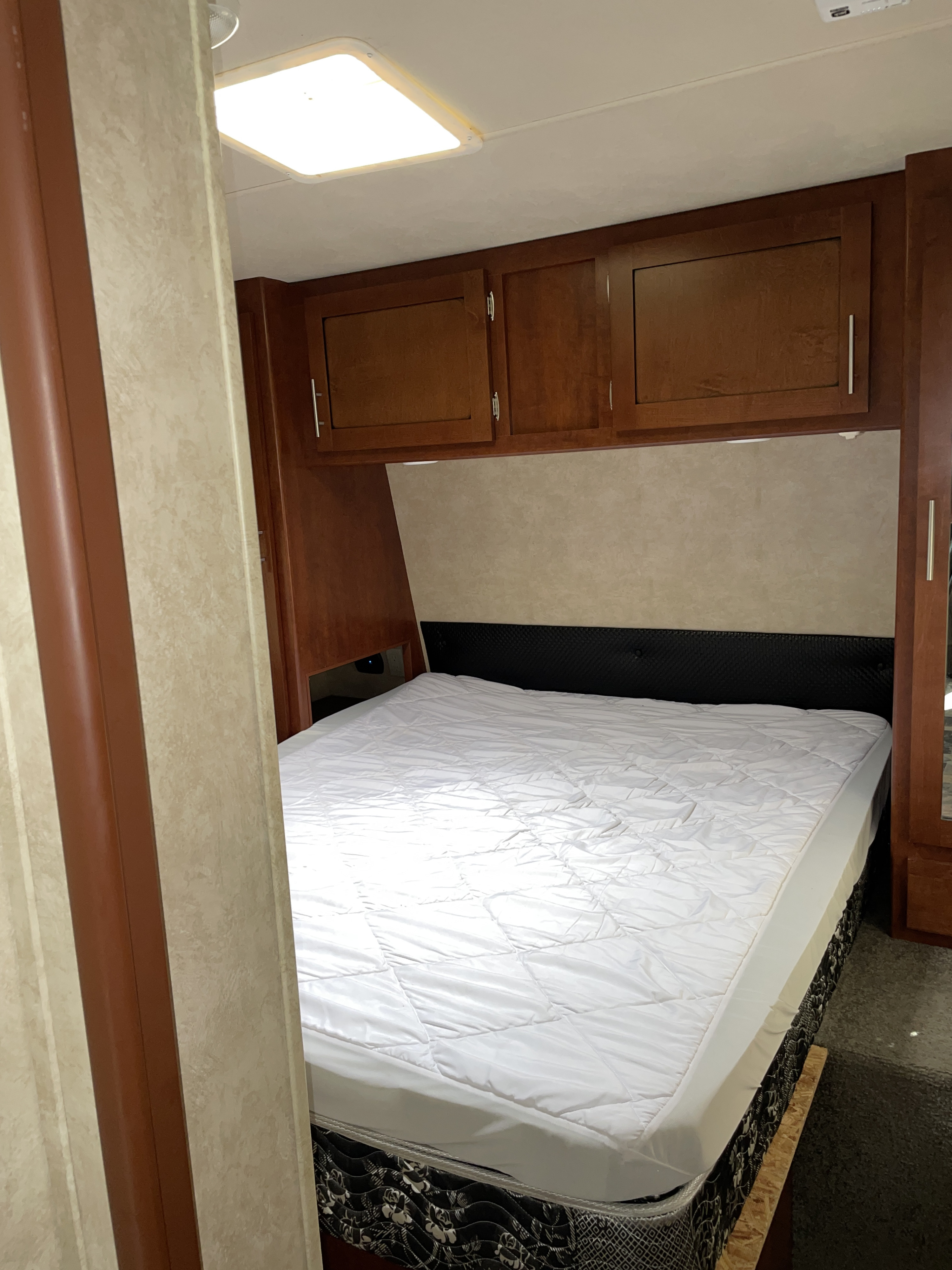 Inside an RV rental, a compact bedroom with a white-quilted bed, black headboard, and wood overhead cabinets.. Forest River Shockwave 2014