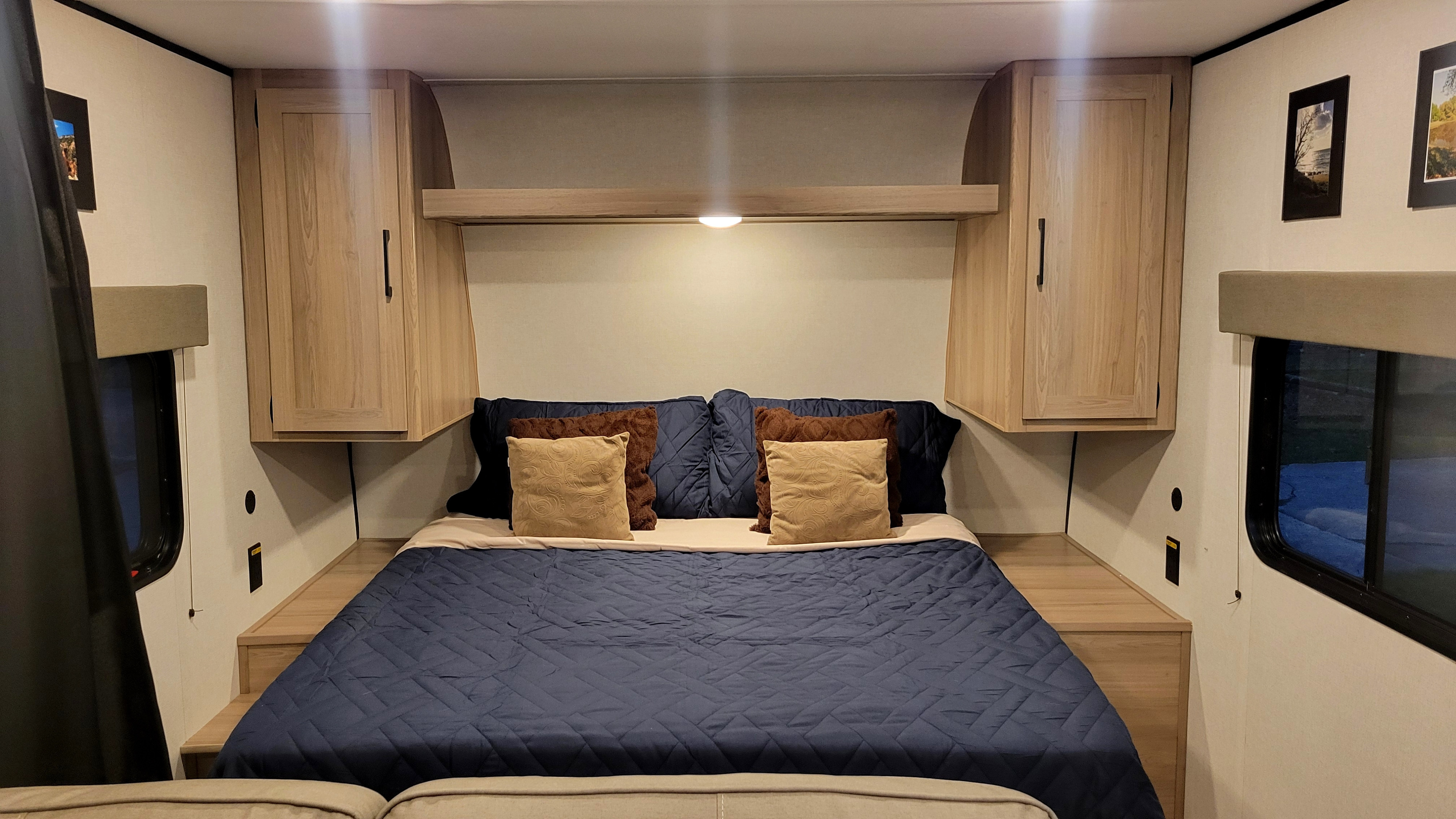 Explore this RV rental's inviting bedroom, featuring a comfy bed with navy bedding, overhead cabinets, and bright windows.. Coleman 23B 2025