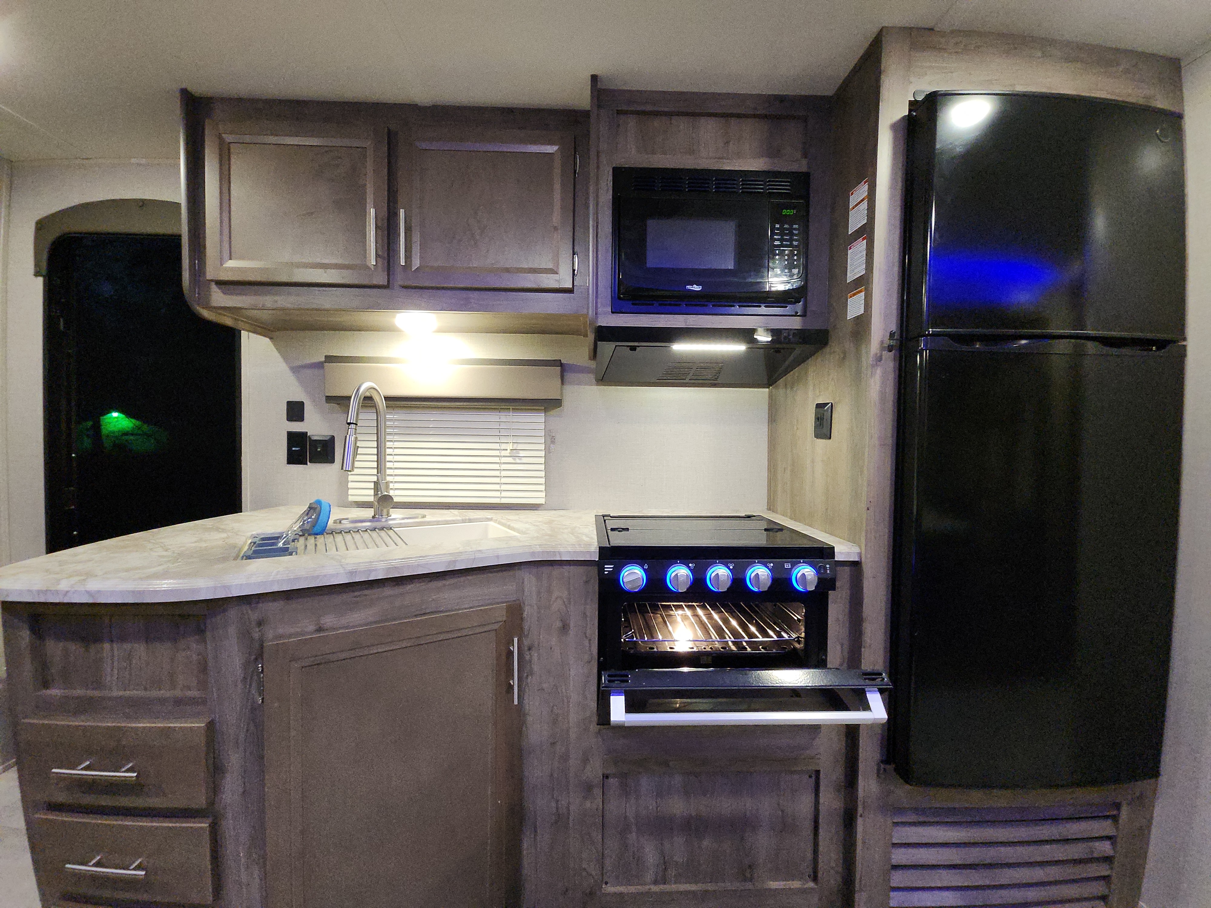 Inside an rv rental: a compact kitchen with a stove, microwave, large fridge, sink, and wood-toned cabinets. Blue stove lights add detail.. Forest River Aurora Aurora 2021