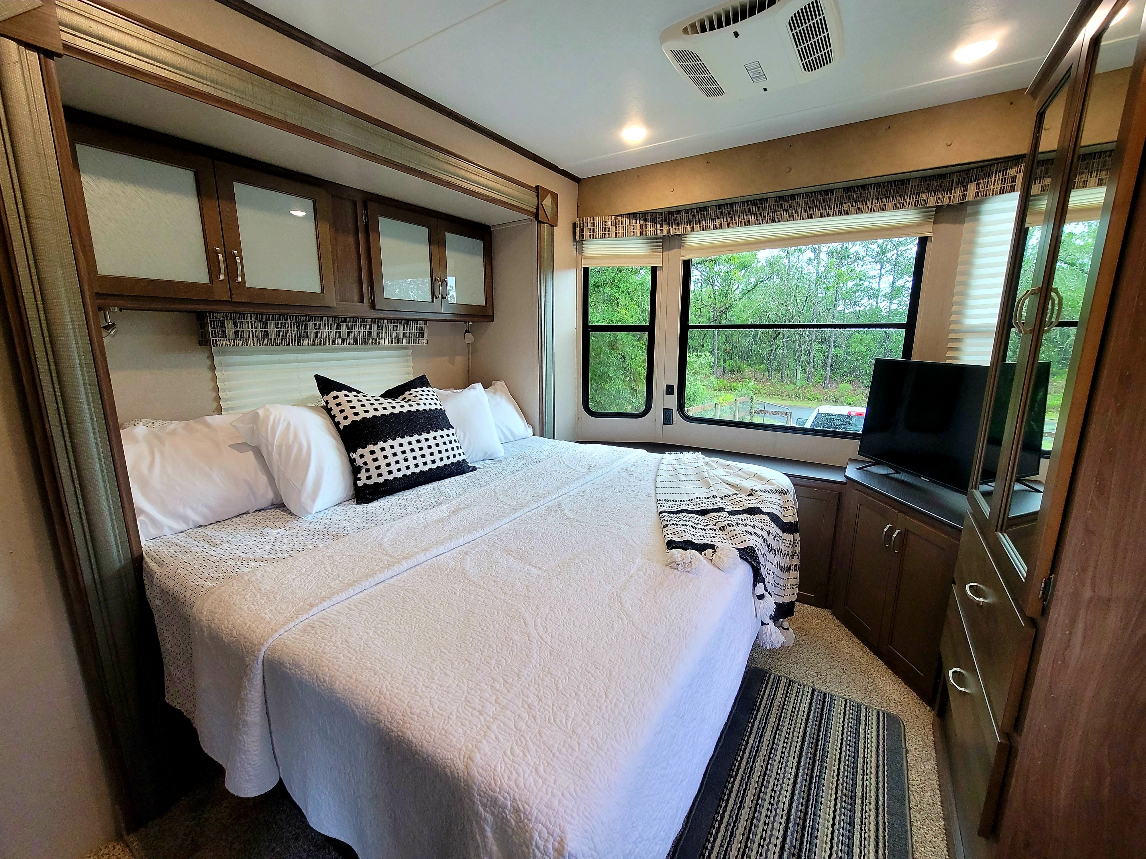 Cozy RV rental bedroom featuring a queen bed with white bedding, large windows with nature views, a TV, and storage cabinets.. Keystone Retreat 2019