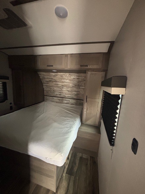 Comfortable RV rental bedroom with a bed, rustic headboard, overhead cabinets, and a window with privacy blinds.. Gulf Stream Amerilite 2023