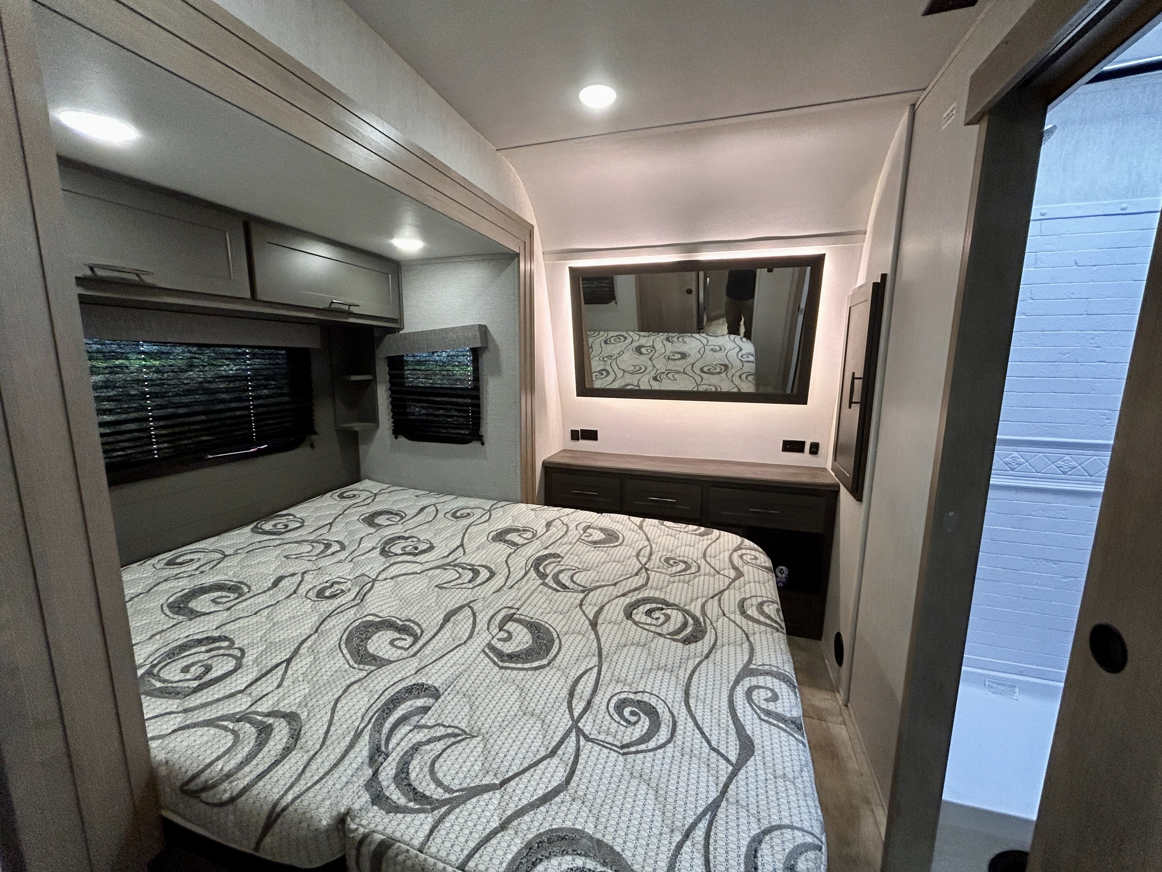 Spacious and modern RV rental bedroom featuring a patterned bed, overhead cabinets, a dresser with mirror, and adjoining bathroom.. Grand Design Momentum 2023