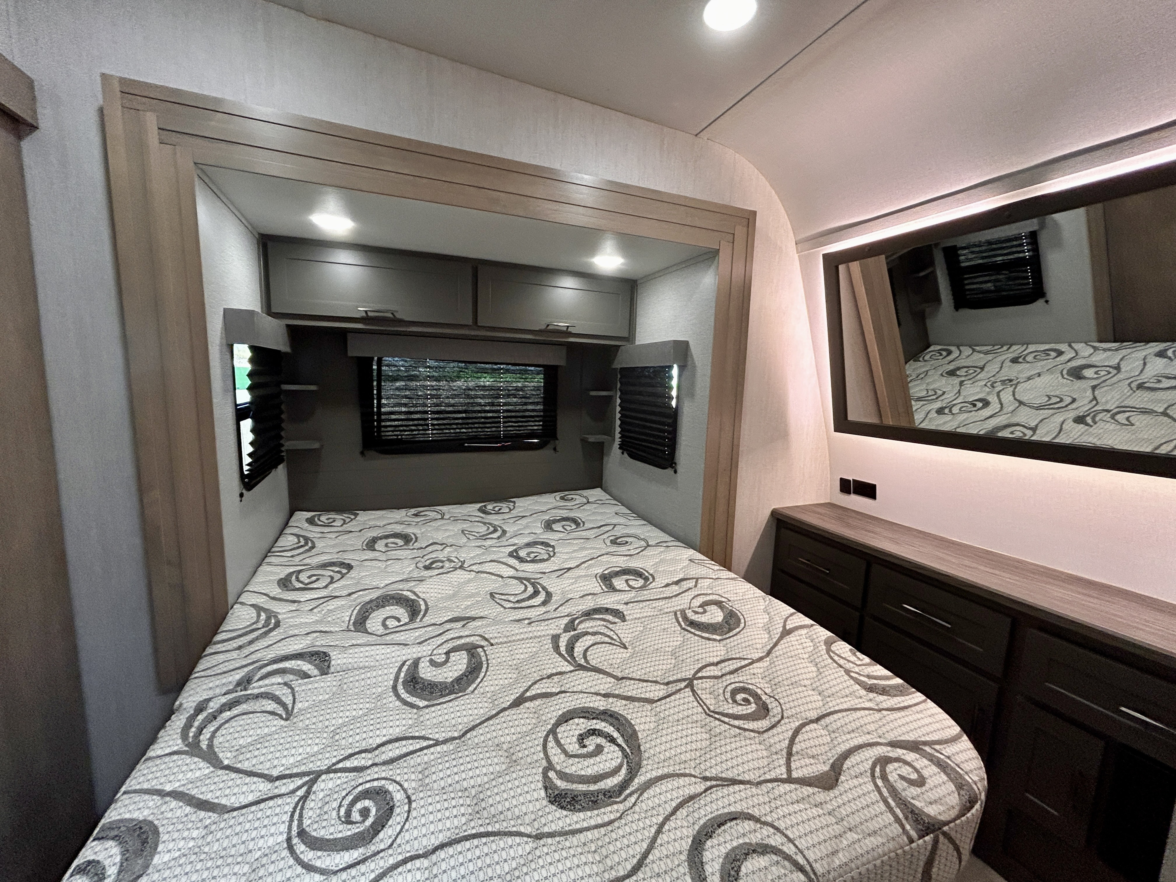 Spacious RV rental bedroom featuring a patterned bed, overhead cabinets, window, dresser, and illuminated mirror.. Grand Design Momentum 2023