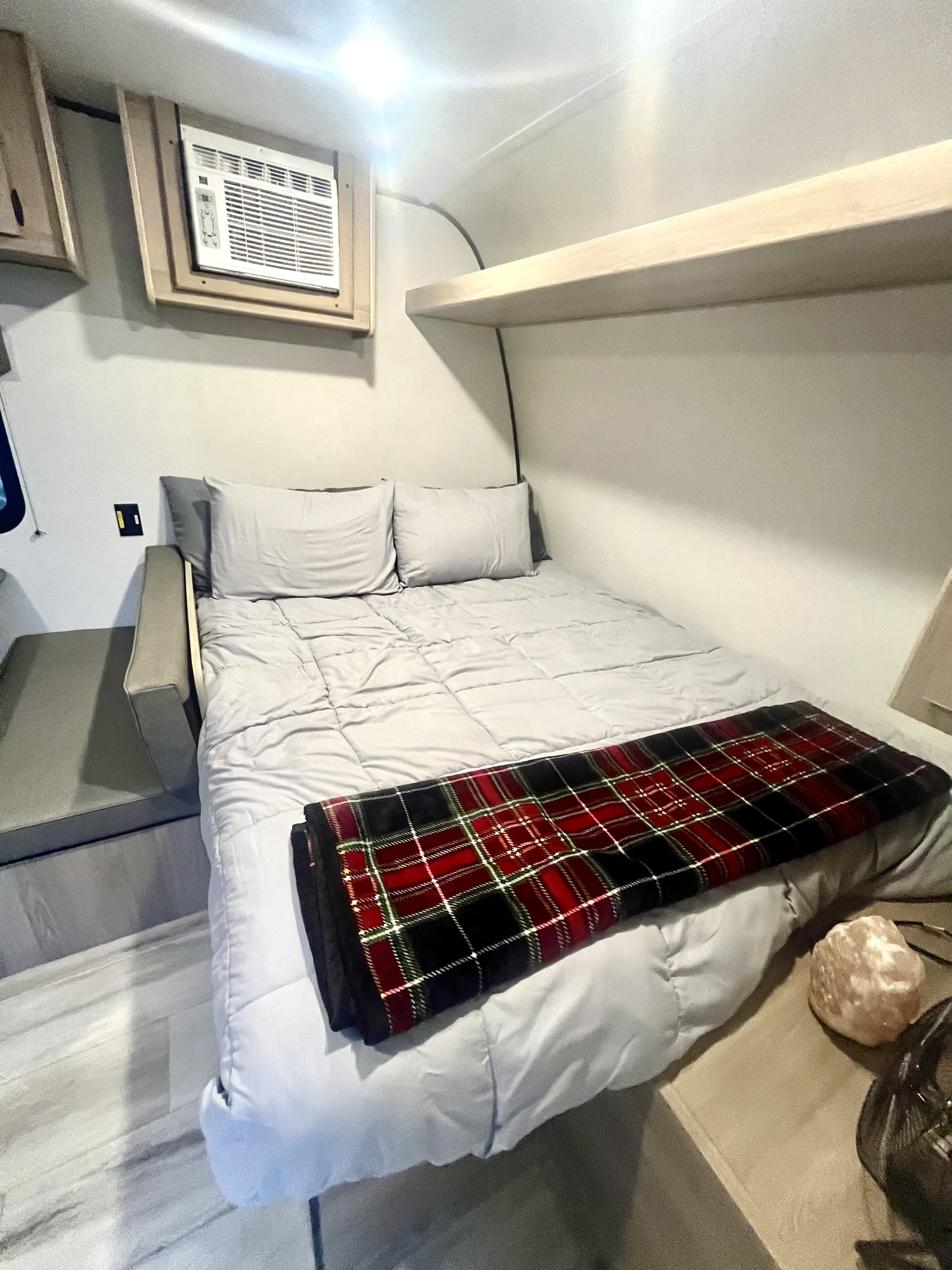 Cozy RV rental interior with a light grey bed, plaid blanket, mounted AC unit, and overhead shelf.. Coleman Other 2025