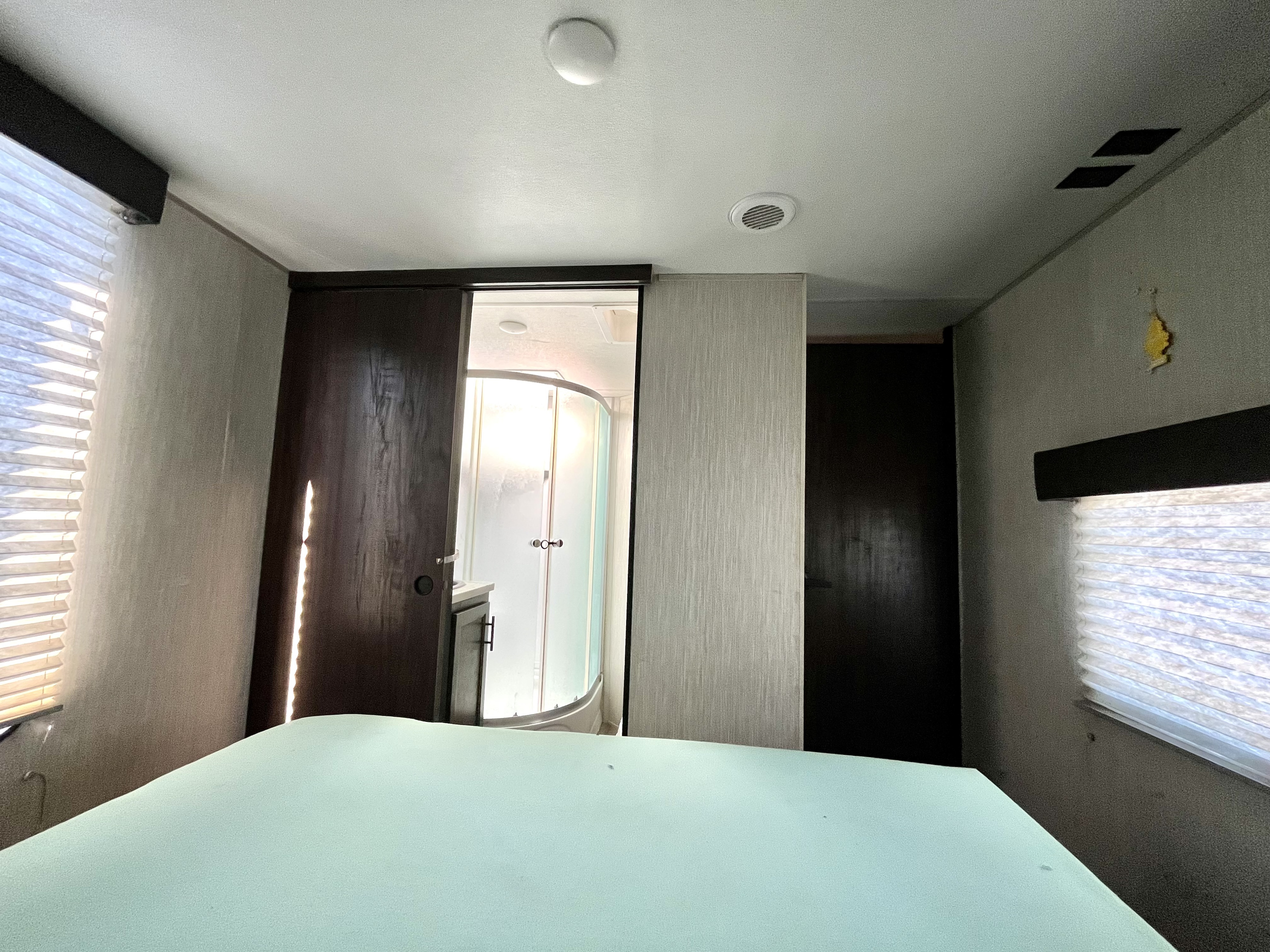 RV rental bedroom with light blue bed, windows/blinds, and sliding door to compact bathroom with curved shower.. Forest River Cherokee 2020