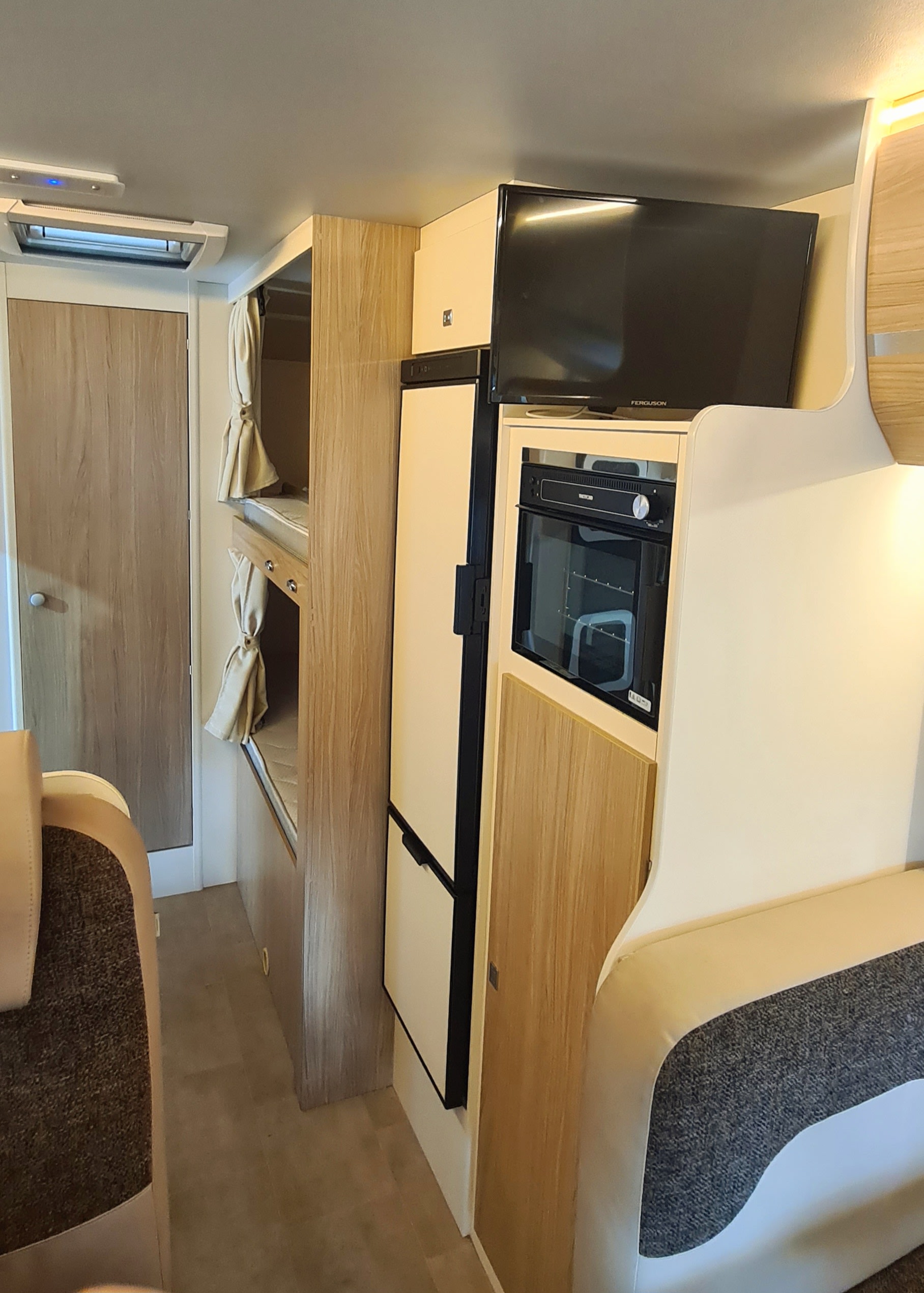 RV rental interior: light wood cabinetry houses bunk beds, a refrigerator, an oven, and a mounted TV in this compact camper.. Ford Transit Rimor Evo Sound 7 2022