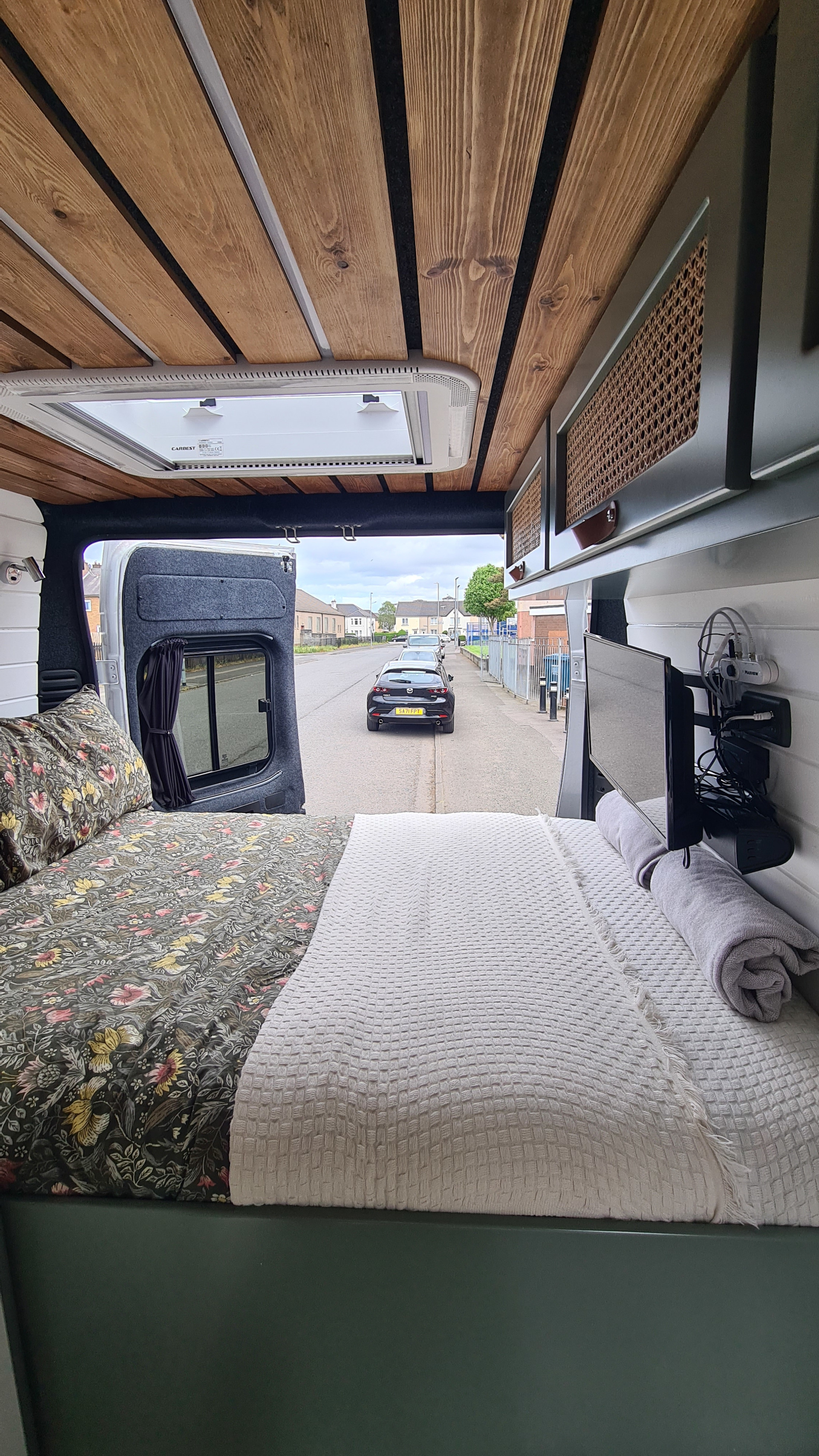Inside a stylish RV rental: comfy bed, wood ceiling, TV, and street view from open rear doors.. Citroen Relay L3H2 2022