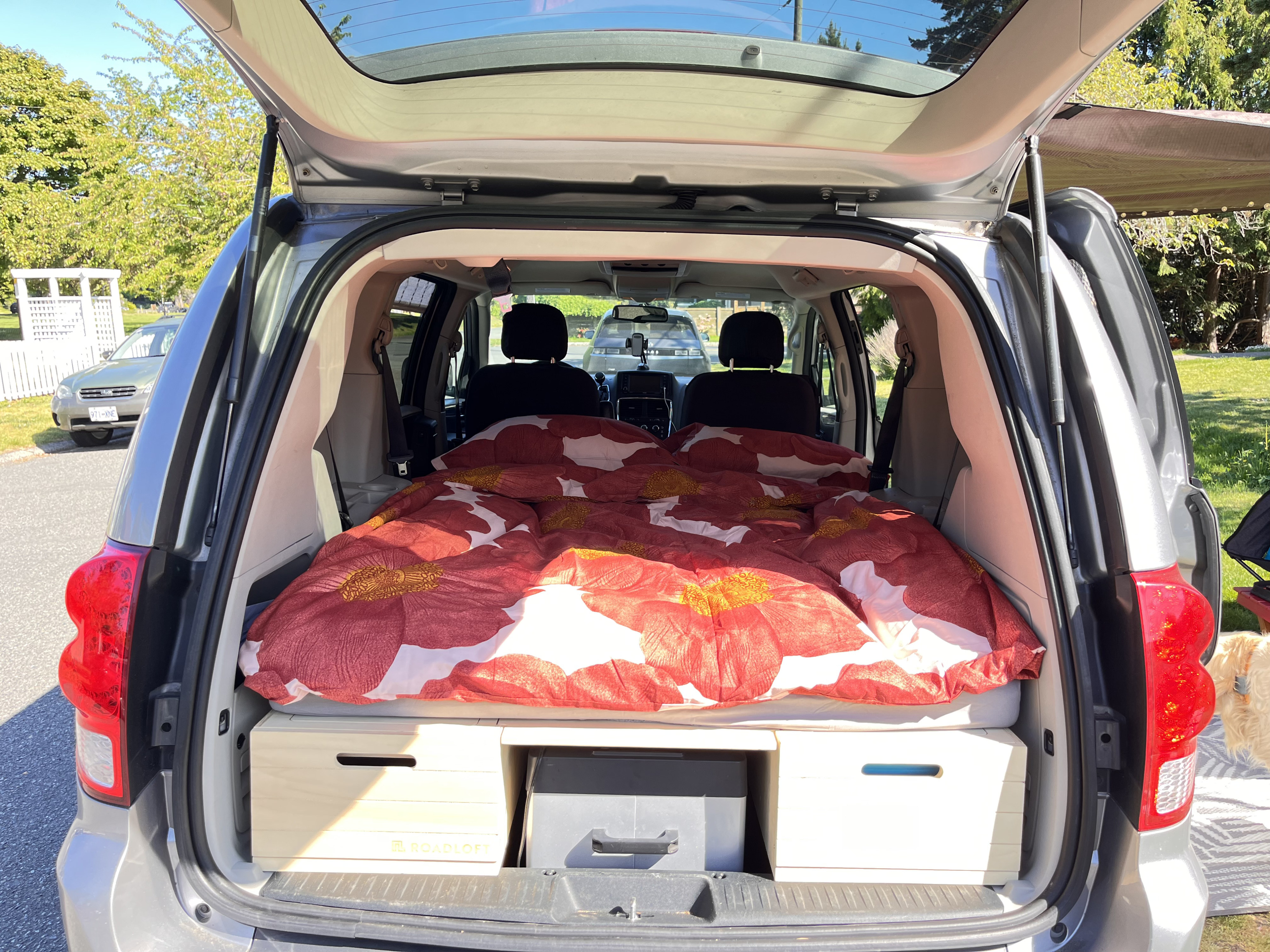Minivan camper setup, tailgate open. Inside, a bed with red floral duvet sits atop storage boxes, ready for your next rv rental adventure.. Dodge Grand Caravan 2013