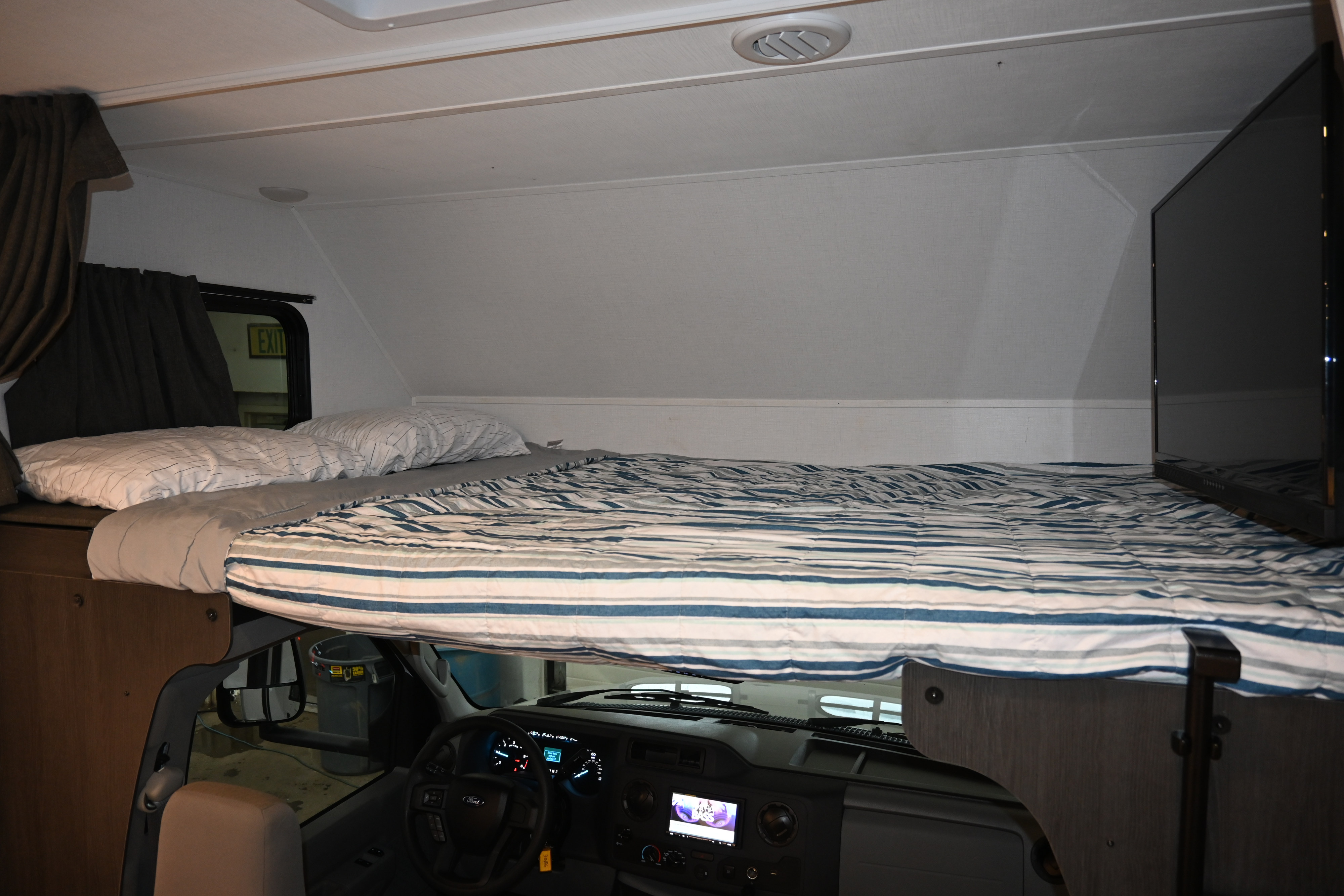 Cozy RV rental cab-over bunk bed with striped comforter, TV, and dashboard view from the sleeping area.. Forest River Sunseeker - LE Series M-2850S 2024