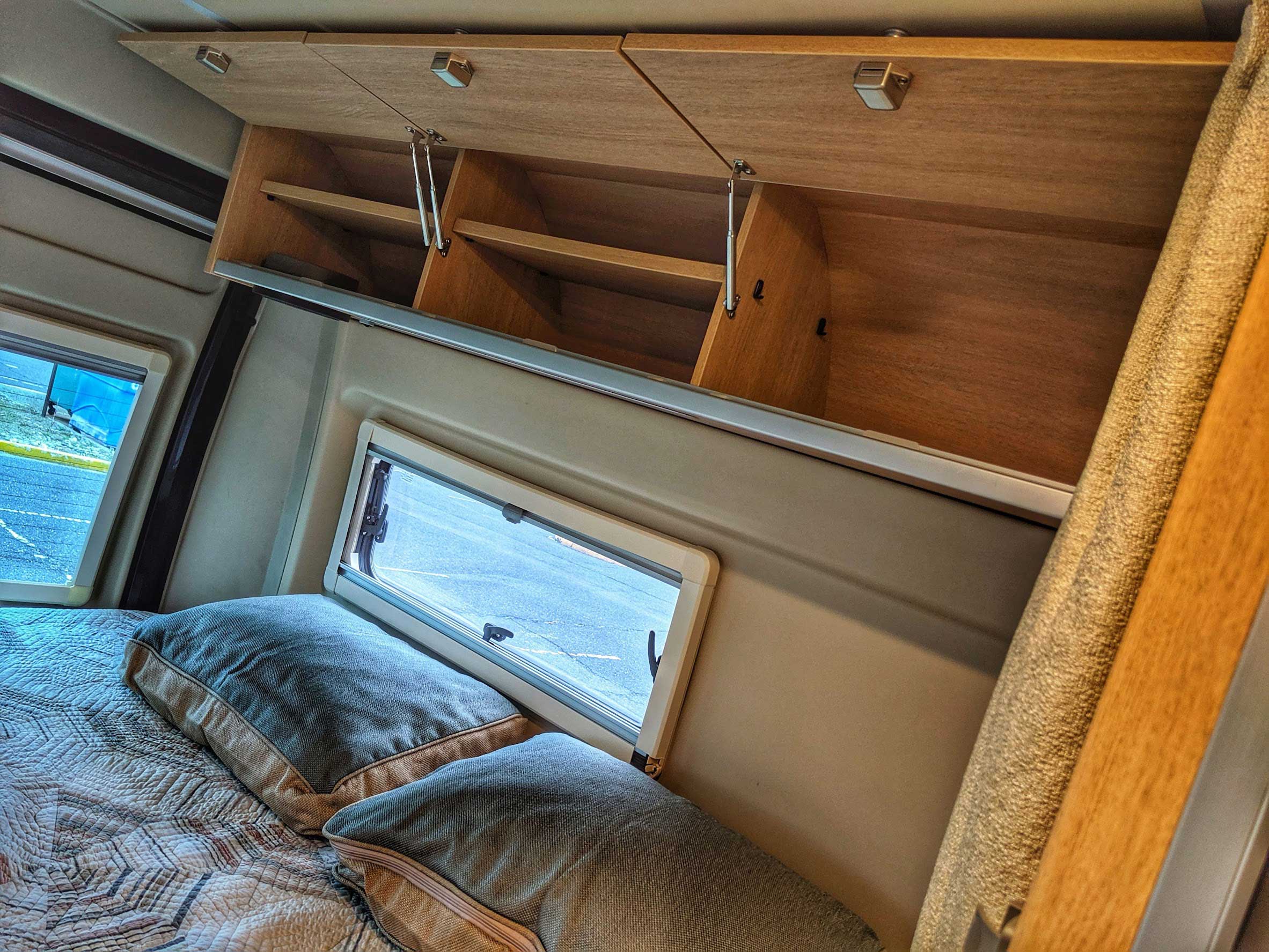RV rental bed area with plush pillows, patterned quilt, open overhead storage cabinets, and windows.. Hymer Aktiv 2.0 2019