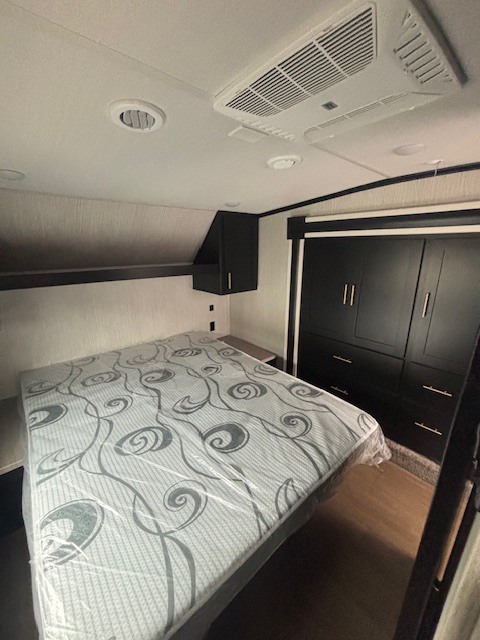 Modern RV rental bedroom featuring a bed with plastic-covered mattress, ceiling AC, and sleek dark storage units.. Forest River Wildcat 2021