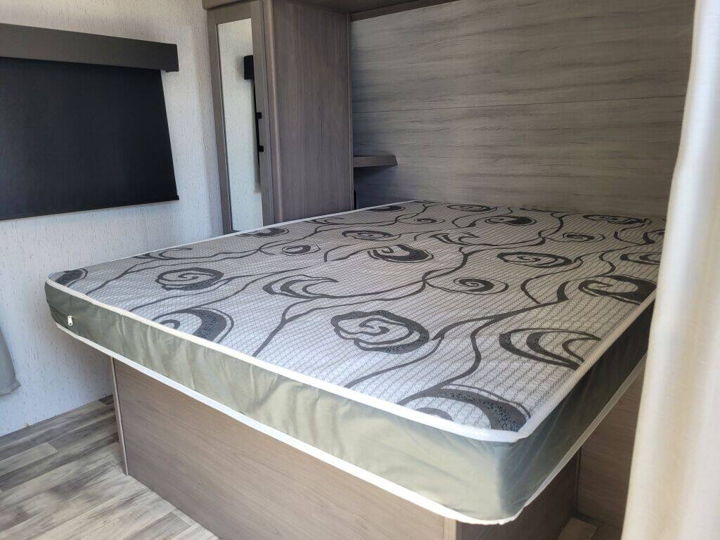 RV rental bed with patterned mattress, wood interior, window blind, and mirrored cabinet.. Keystone Kodiak 2022