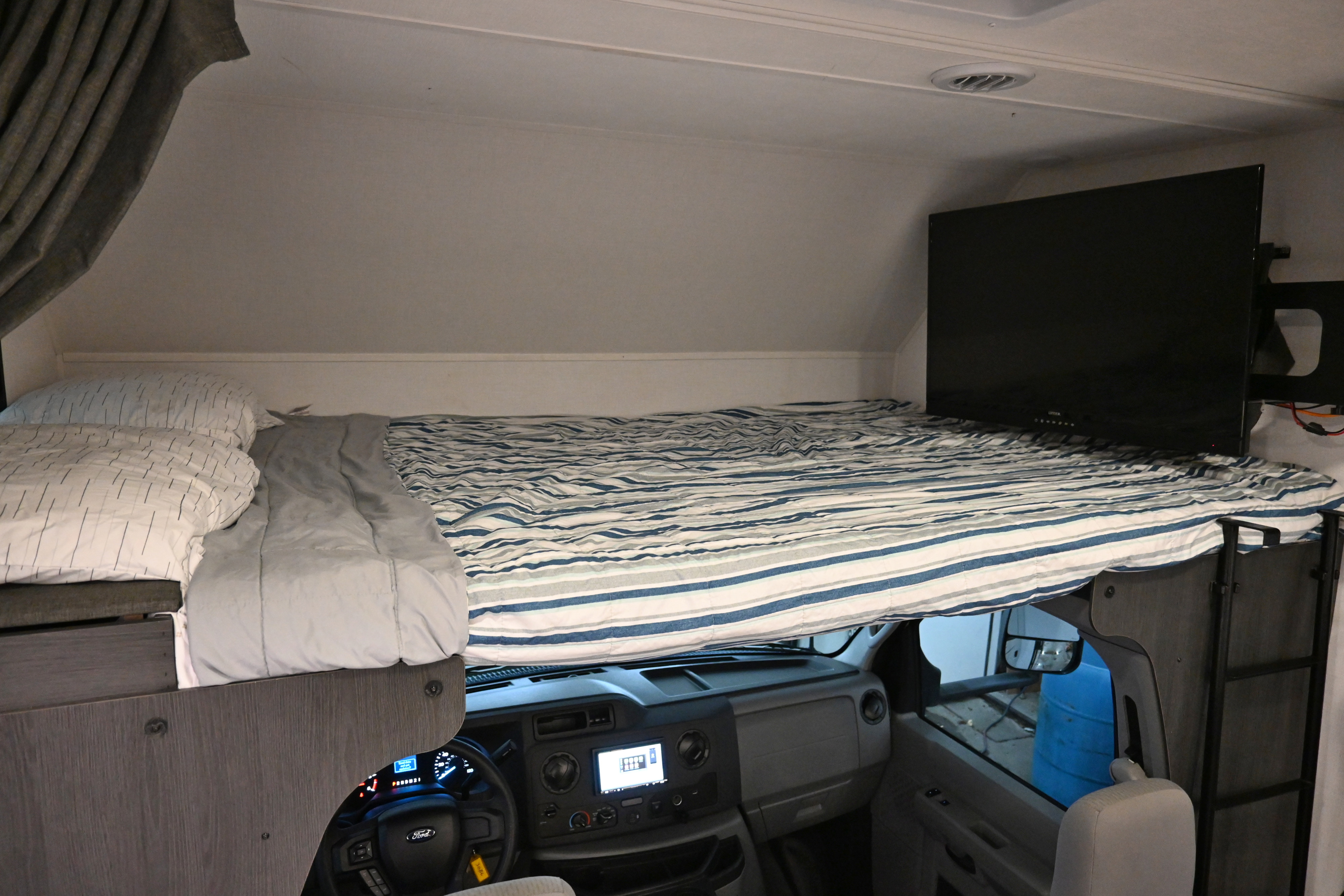 RV rental interior: cab-over bunk bed with striped bedding, TV, and the driver's cockpit below. Ideal for trips.. Forest River Sunseeker - LE Series M-2850S 2024