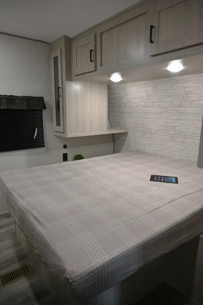 Comfortable RV rental bedroom, featuring light wood overhead cabinets, under-cabinet lighting, and a cozy plaid bed for your trip.. Coachmen Catalina Legacy 2021