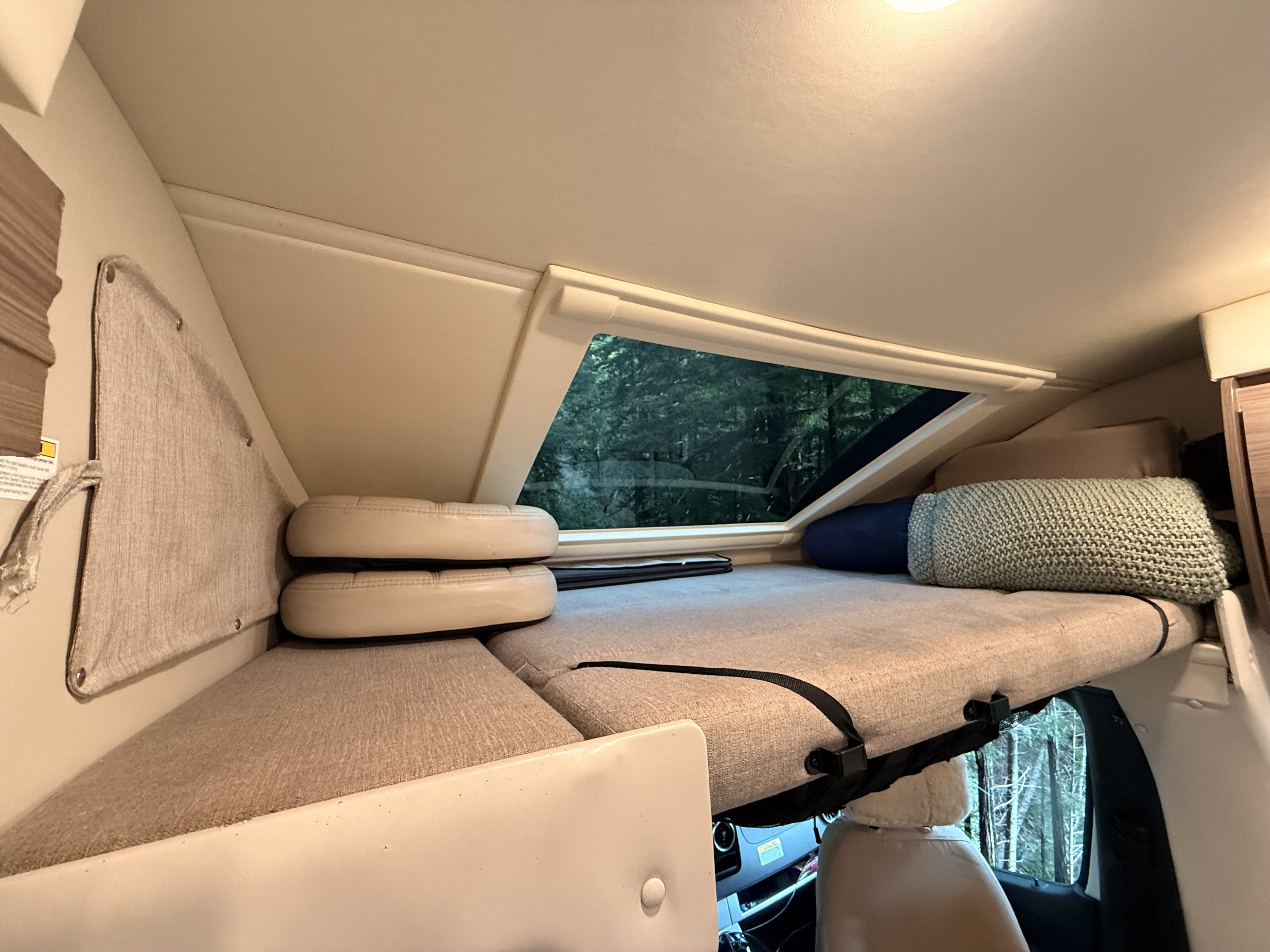 Cozy overhead bunk in an RV rental, featuring a large window with a serene forest view. Complete with cushions and blankets.. Thor Delano Sprinter 2020