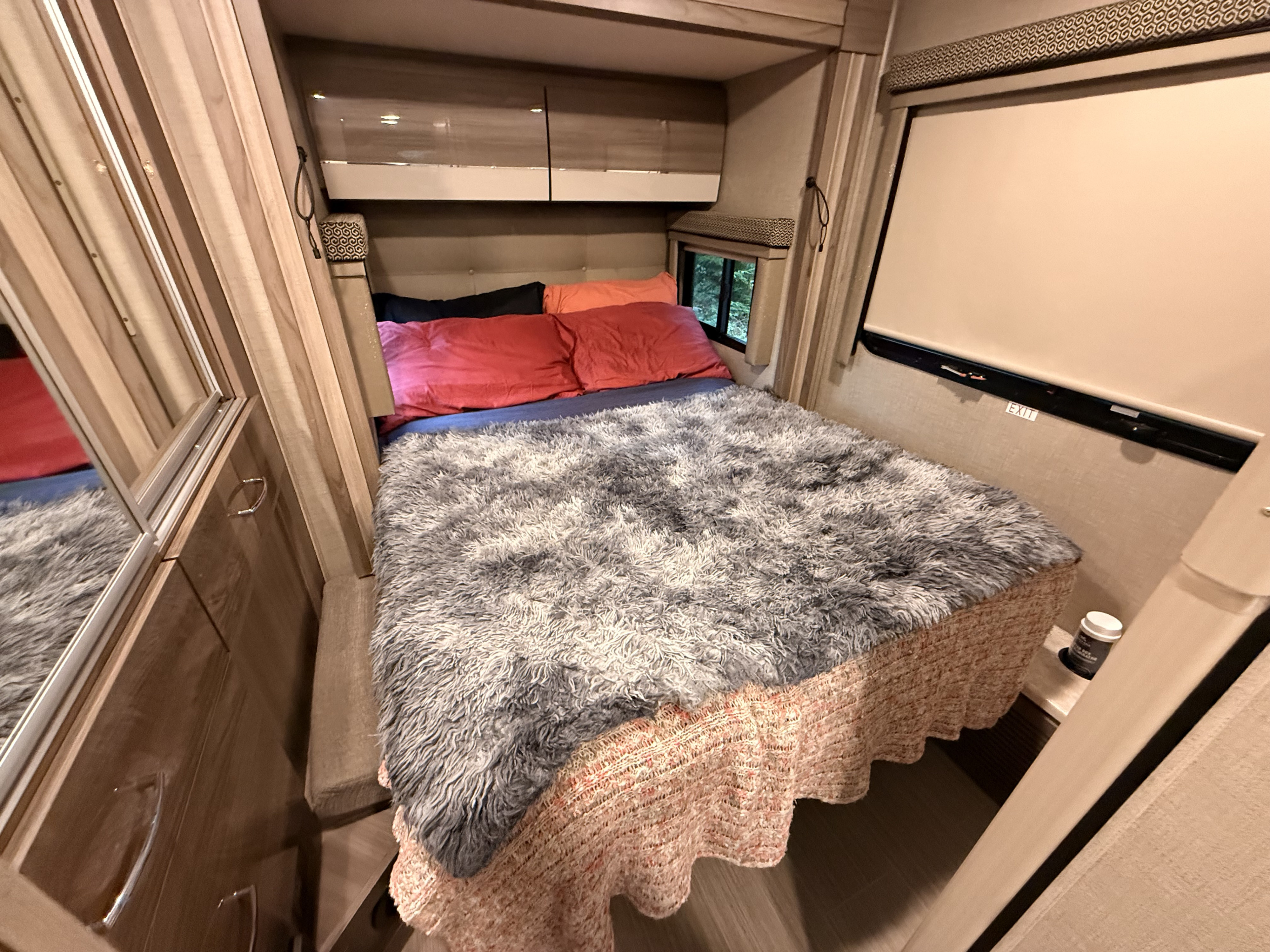 Inside this cozy RV rental bedroom, a large bed with a fluffy grey blanket, colorful pillows, overhead cabinets, and windows.. Thor Delano Sprinter 2020