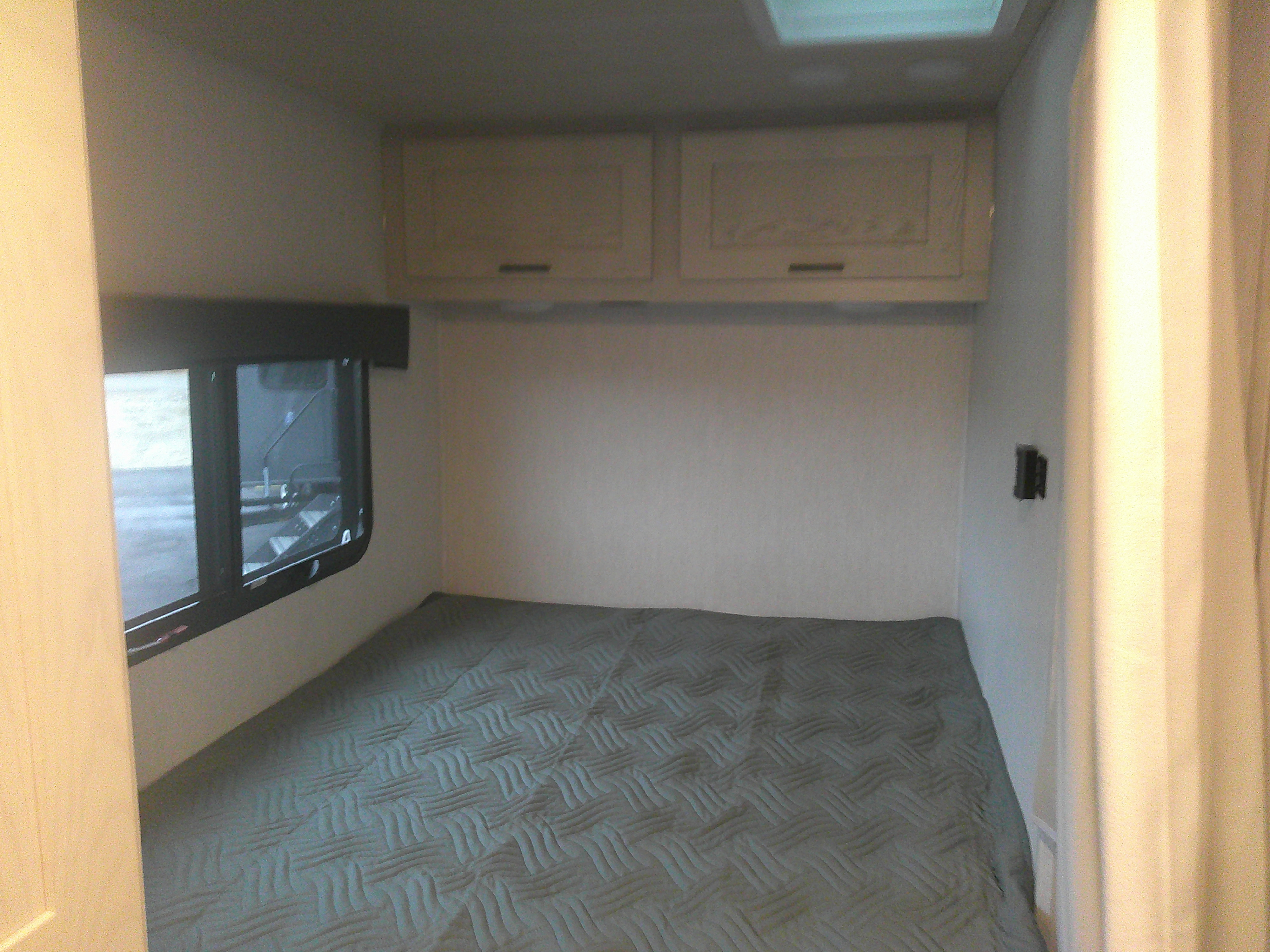 View inside an RV rental bedroom, showing a patterned bed, window, and overhead storage cabinets.. Nexus Phantom 2025