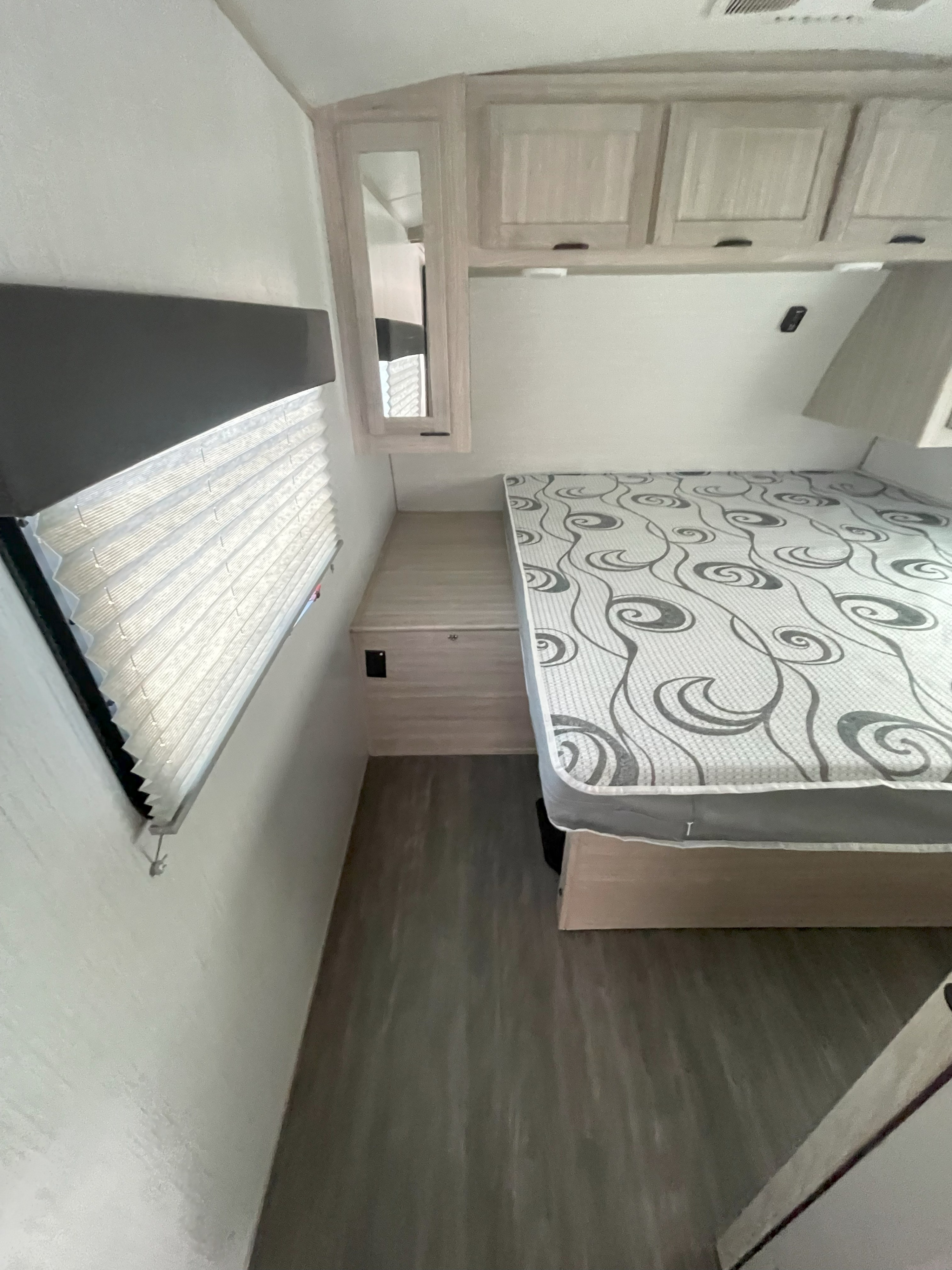 Cozy RV rental bedroom featuring a patterned bed, window with blinds, and light wood storage cabinets.. Cruiser RV Shadow Cruiser 2023