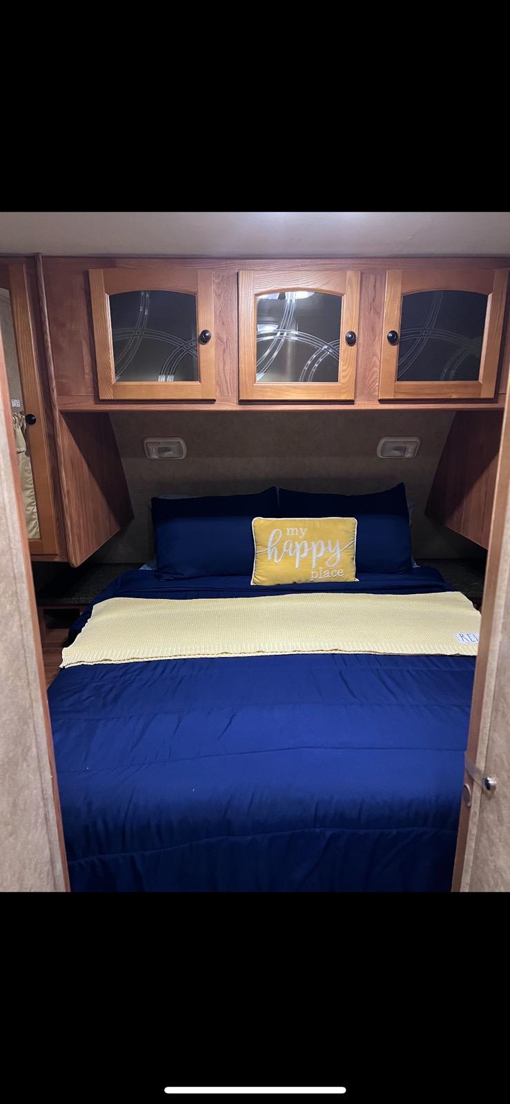 Cozy RV rental bedroom featuring blue bedding, a yellow "my happy place" pillow, and overhead wooden cabinets.. Heartland RVs North Trail 2011