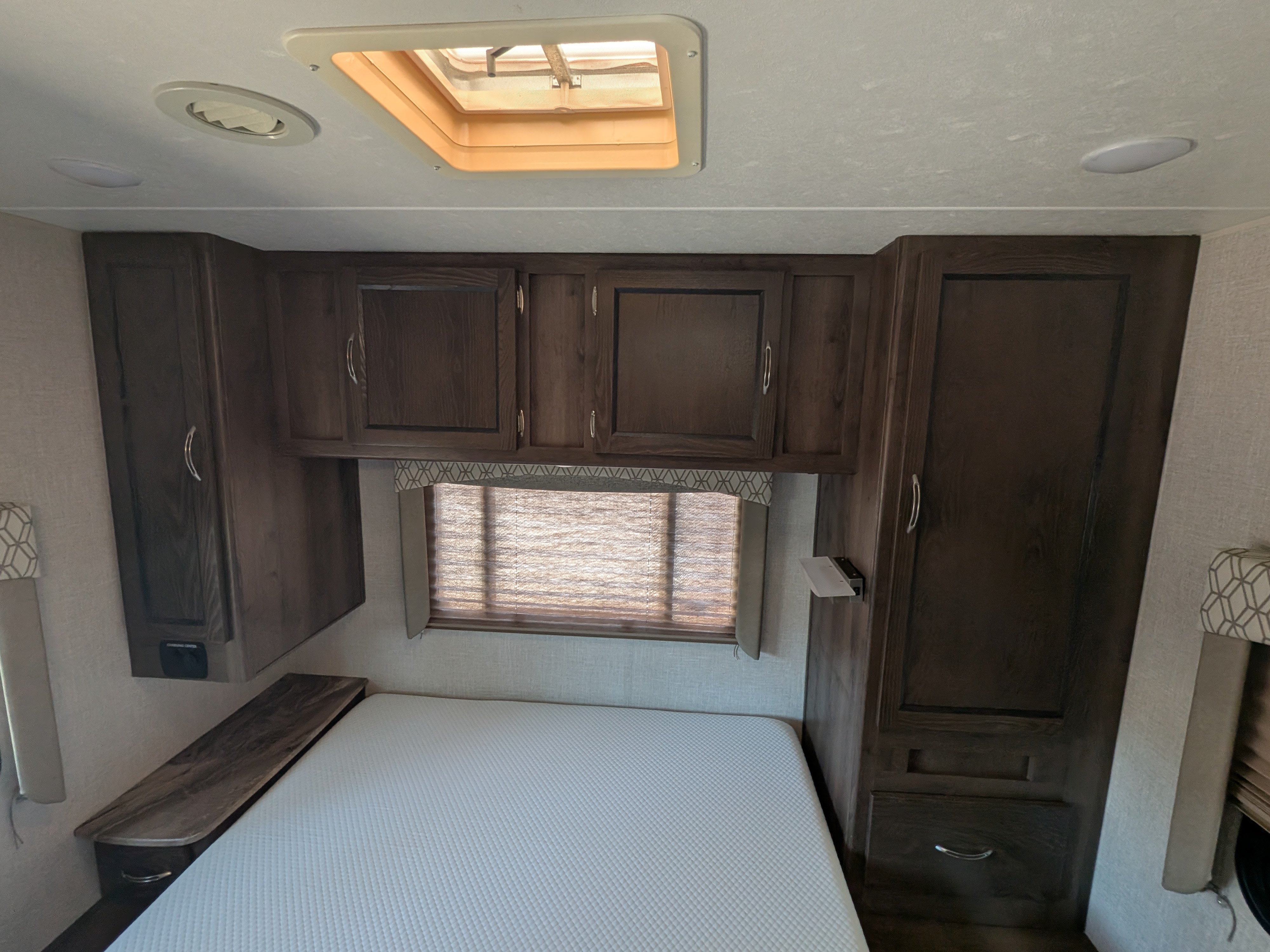 RV rental bedroom featuring a white bed, dark wood overhead cabinets, a window, and ceiling lights/vent for comfortable stays.. Coachmen Freelander 2018