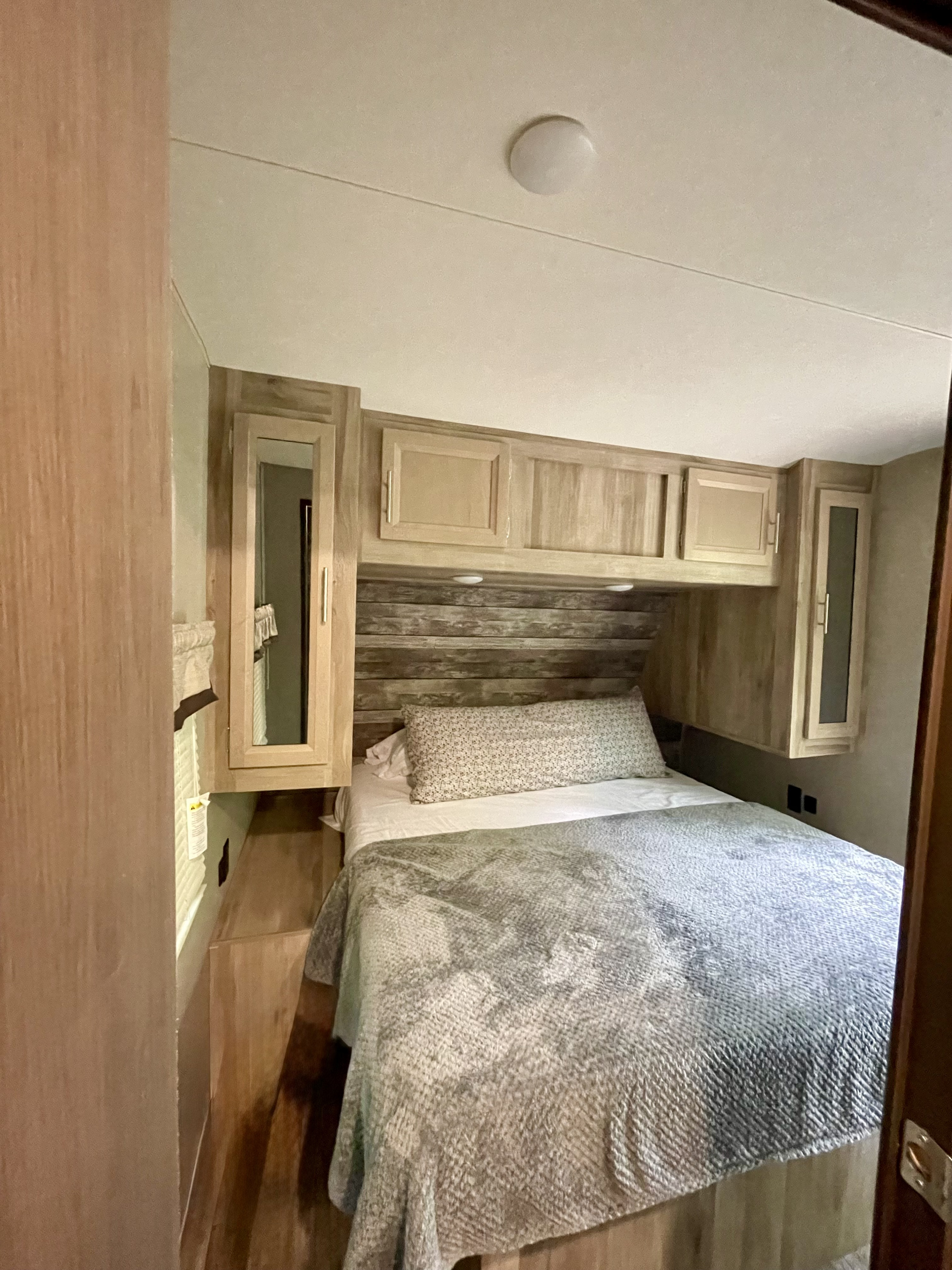 View of a cozy RV rental bedroom with a bed, rustic wood-paneled headboard, and functional storage cabinets.. Coachmen Catalina Legacy Edition 2021