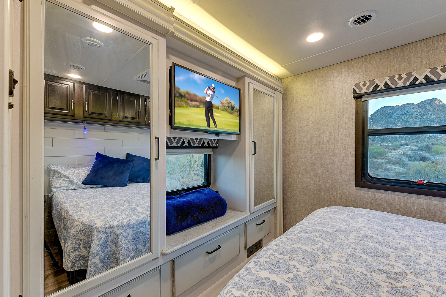 Inside an RV rental bedroom: two beds, TV, mirrored storage, and a window framing desert mountains.. Entegra Coach Vision XL 2023