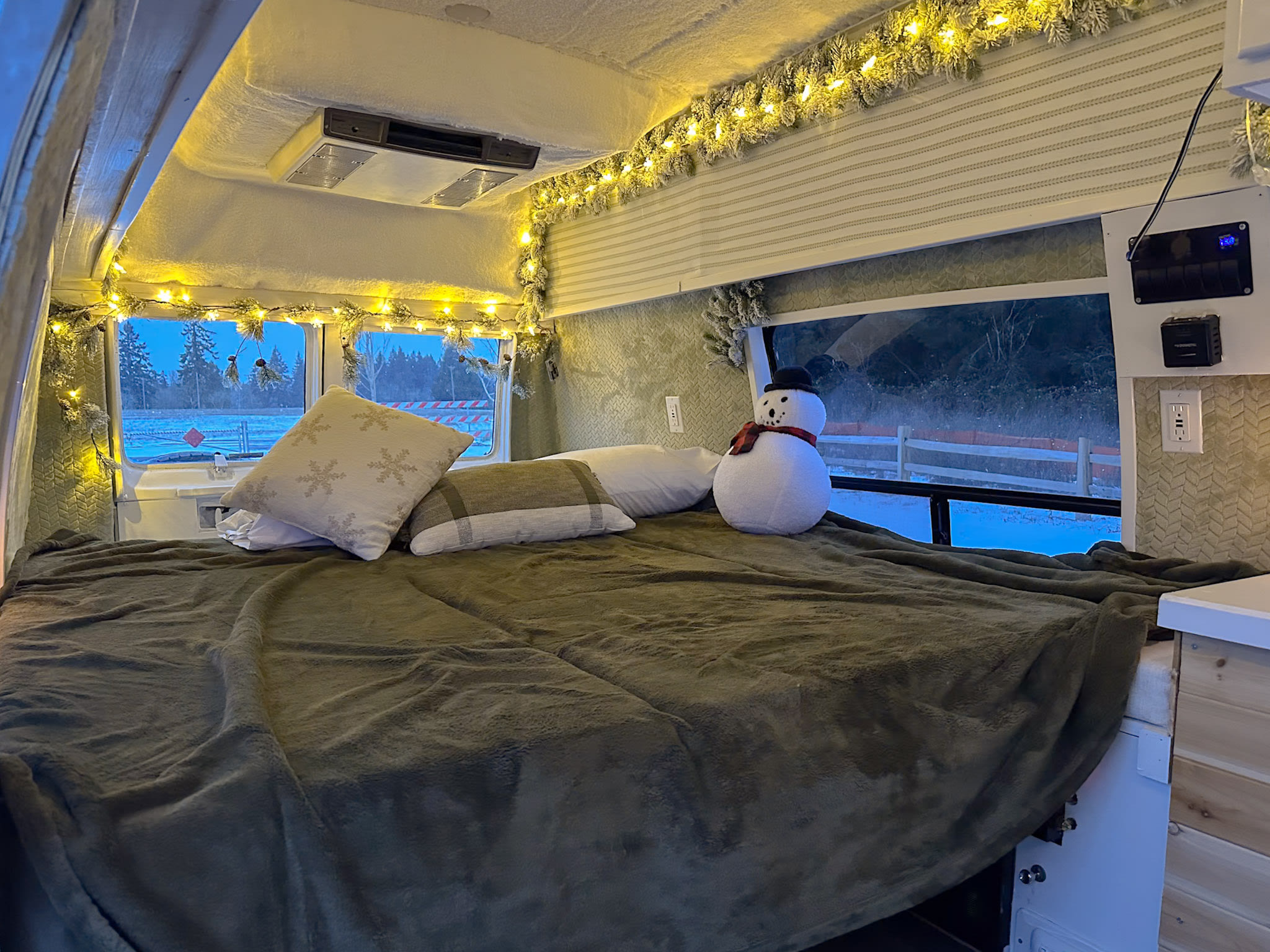 Inside a festive RV rental: a cozy bedroom with warm string lights, snow-flocked garland, soft blankets, and a snowman doll.. Jayco Conversion Van 1998