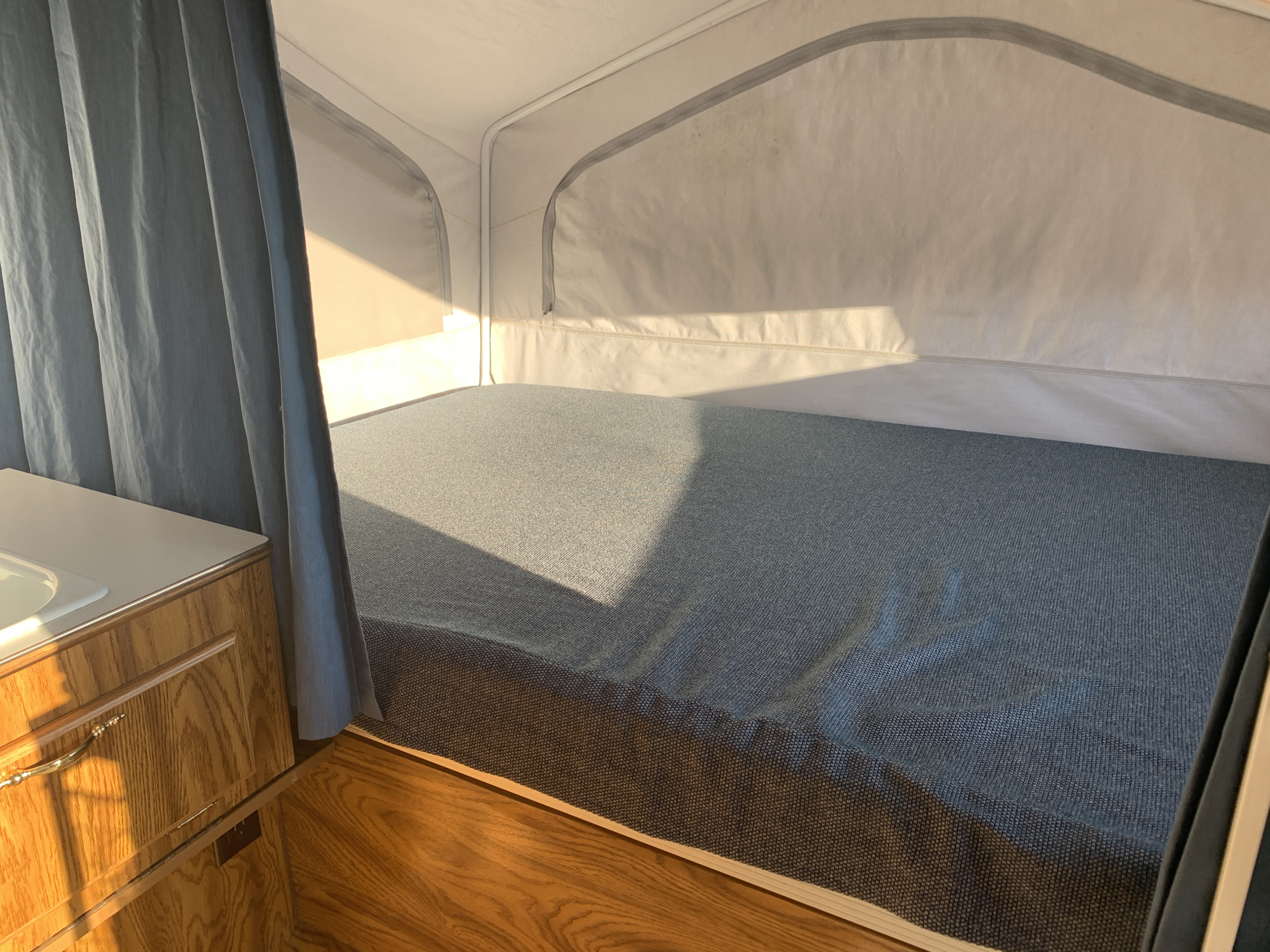 Sunlit pop-up RV rental interior with a bed, blue bedding, white canvas walls, and wood cabinetry.. Forest River Flagstaff 2000