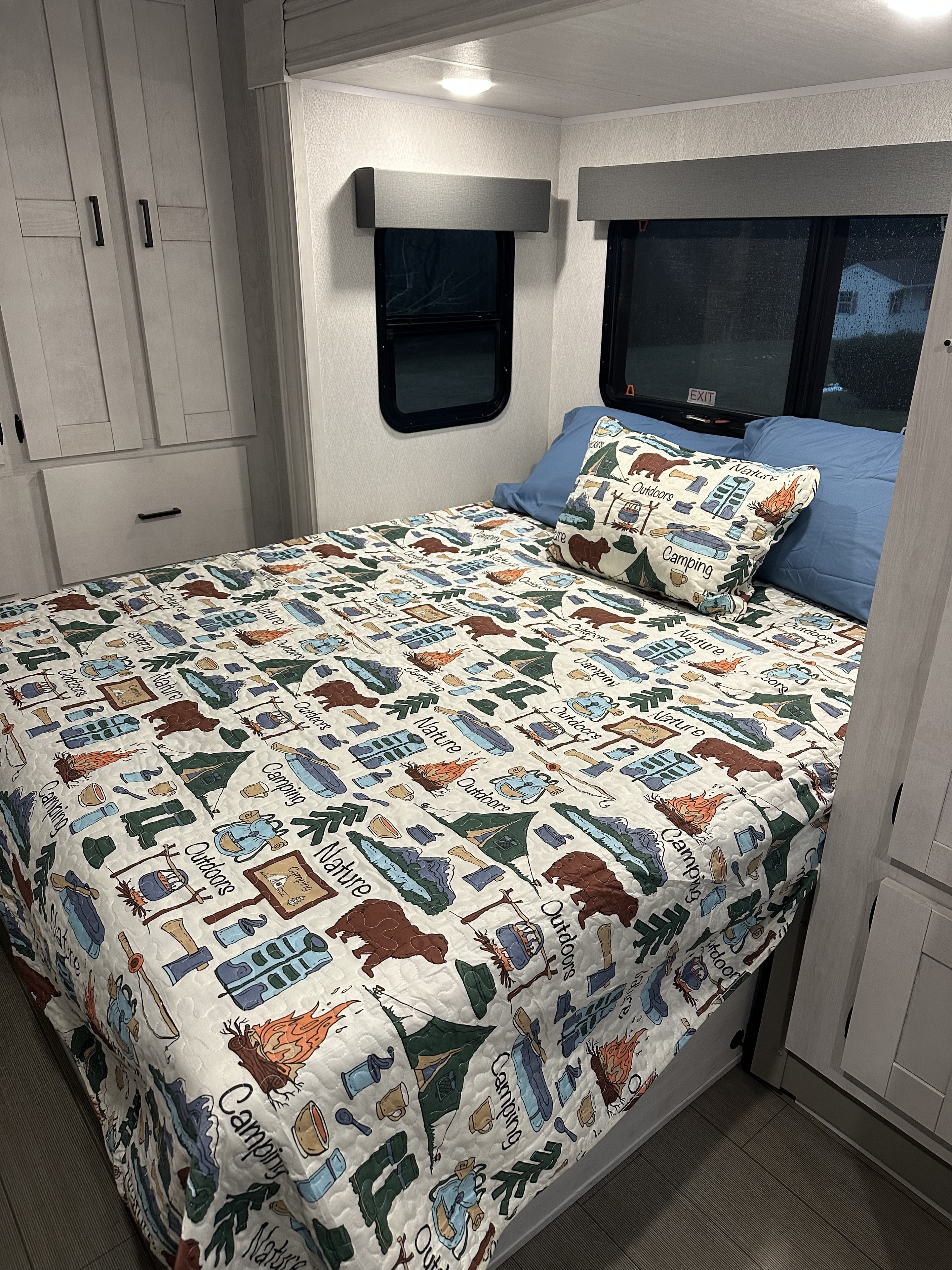 This cozy RV rental bedroom features a camping-themed bedspread, blue pillows, multiple windows, and light gray cabinets, ideal for your next outdoor adventure.. East to West Entrada 24FM 2024