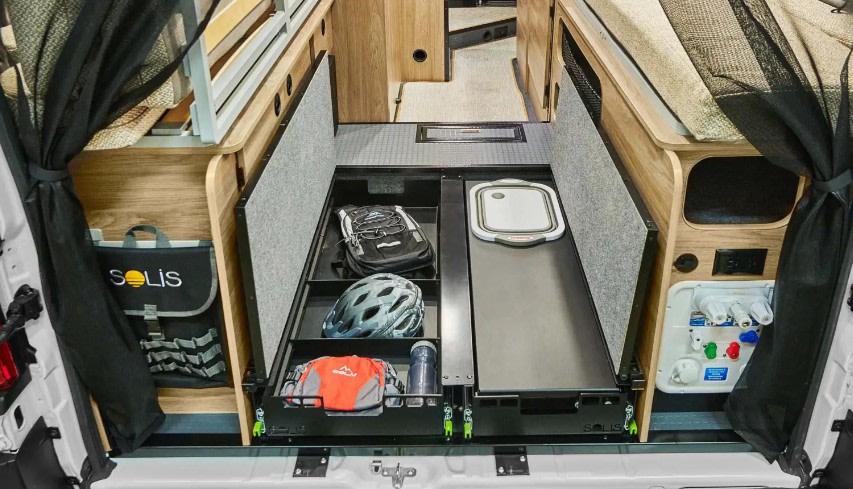 RV rental's organized rear storage area: pull-out drawers hold gear like a backpack & helmet, with water hookups visible.. Winnebago Solis NPF 2025