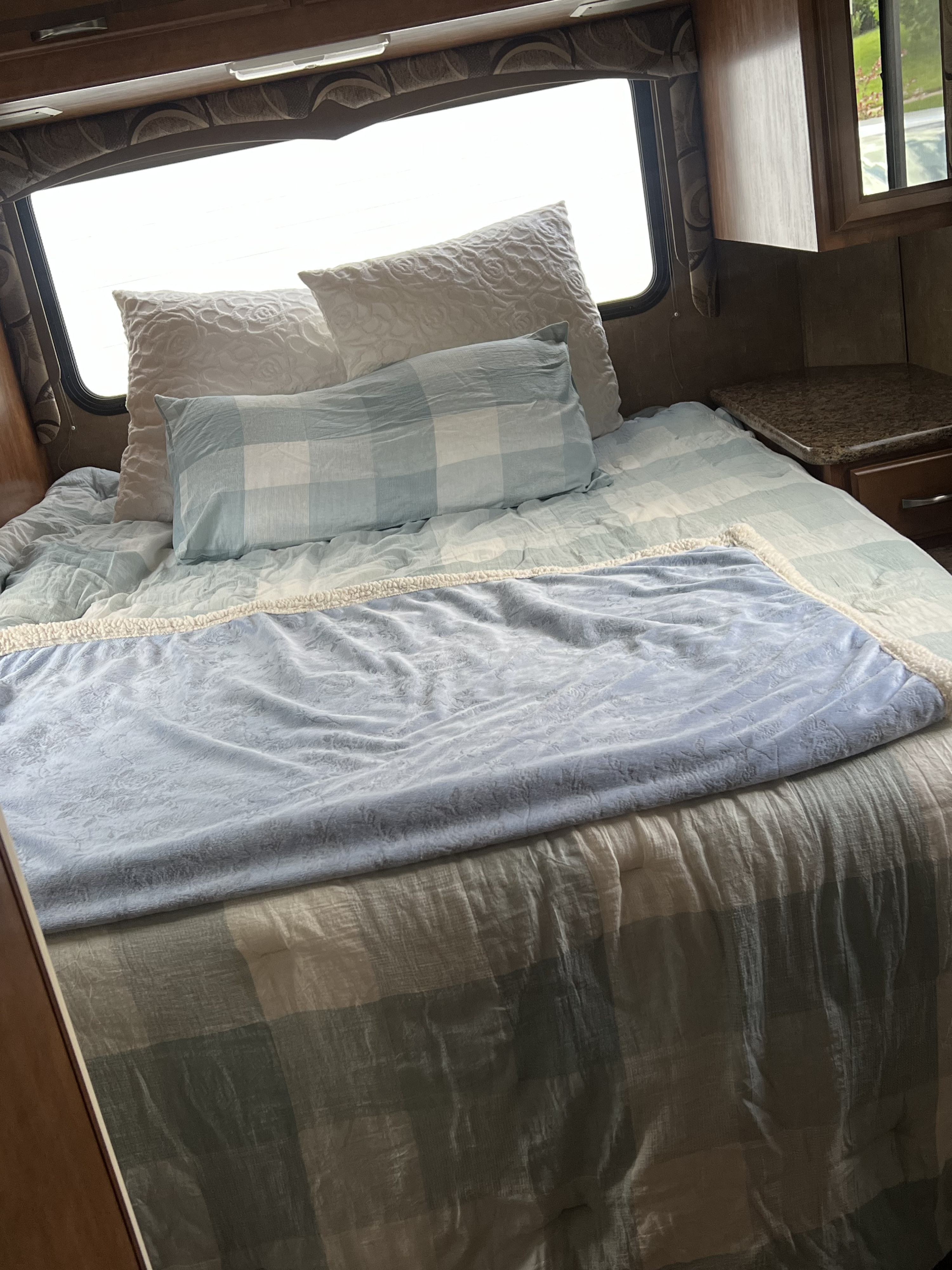 Cozy RV rental bedroom featuring a bed with blue and white plaid bedding, pillows, and a light blue blanket. Includes window and cabinets.. Thor Four Winds 2015