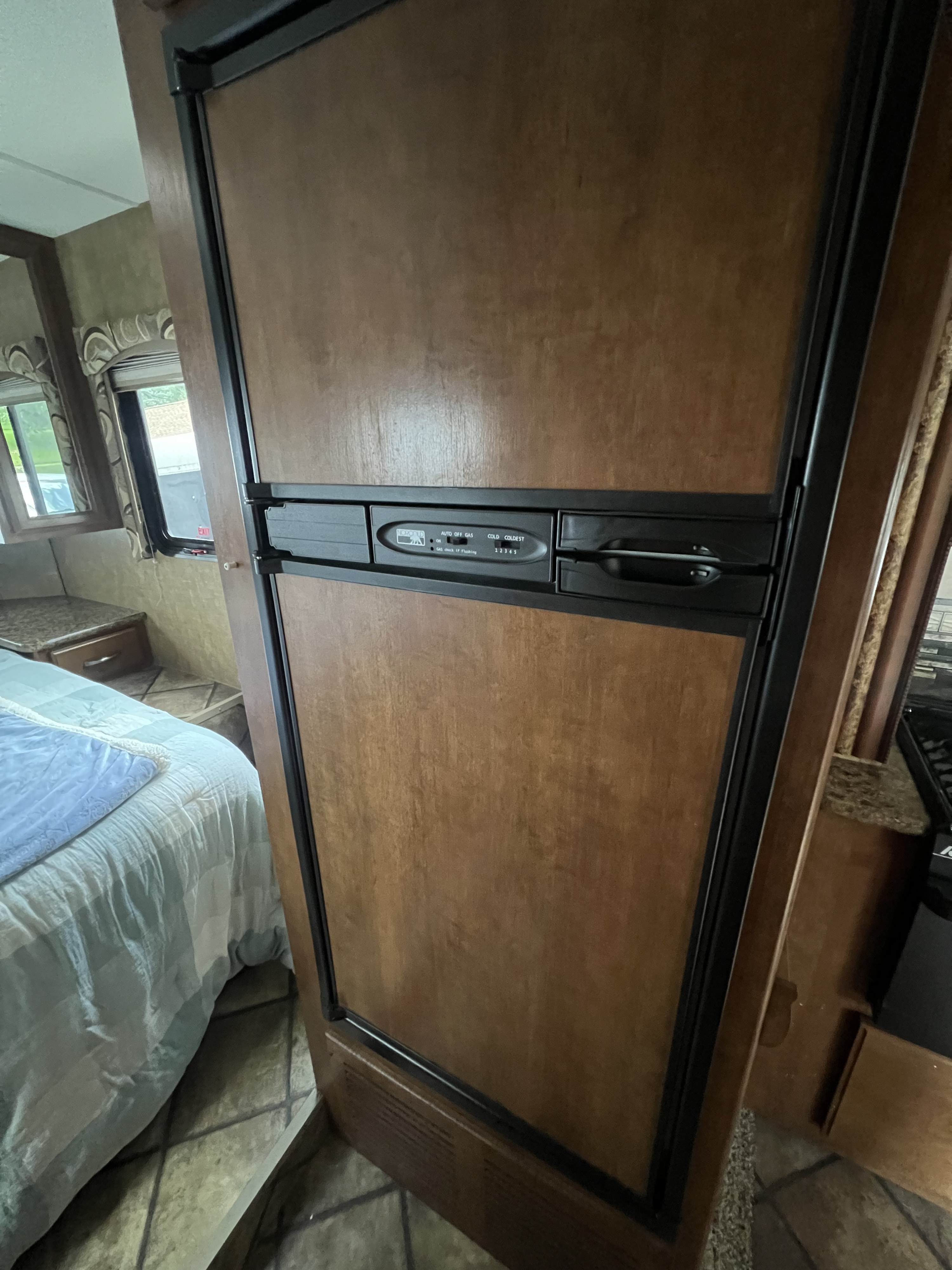 An RV rental interior featuring a wood-paneled refrigerator with controls, alongside a bed and window in a camper.. Thor Four Winds 2015