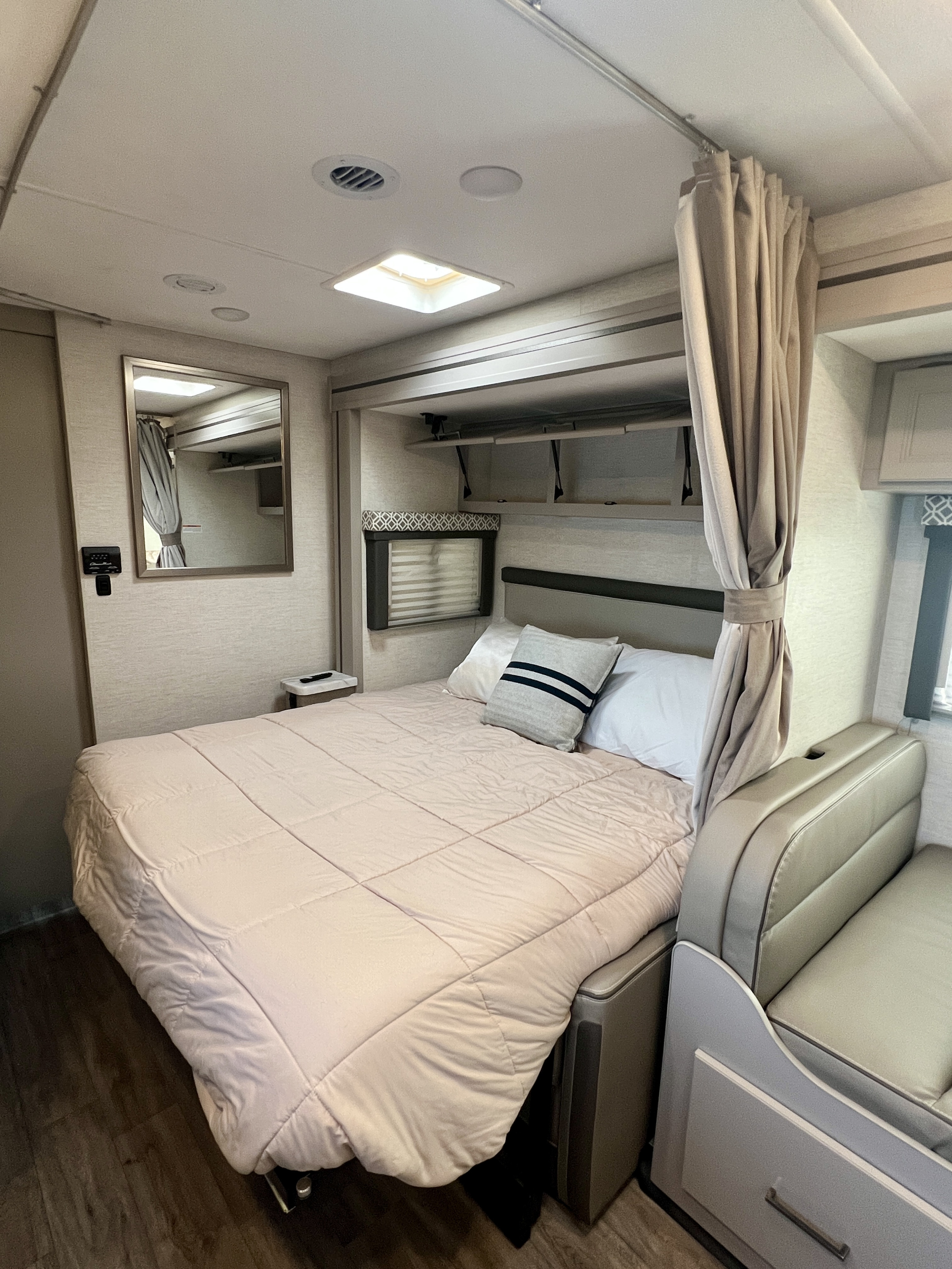 Cozy RV rental bedroom showing a bed, overhead cabinets, a mirror, and attached seating. Ideal for comfortable travel.. Thor Coleman 2024