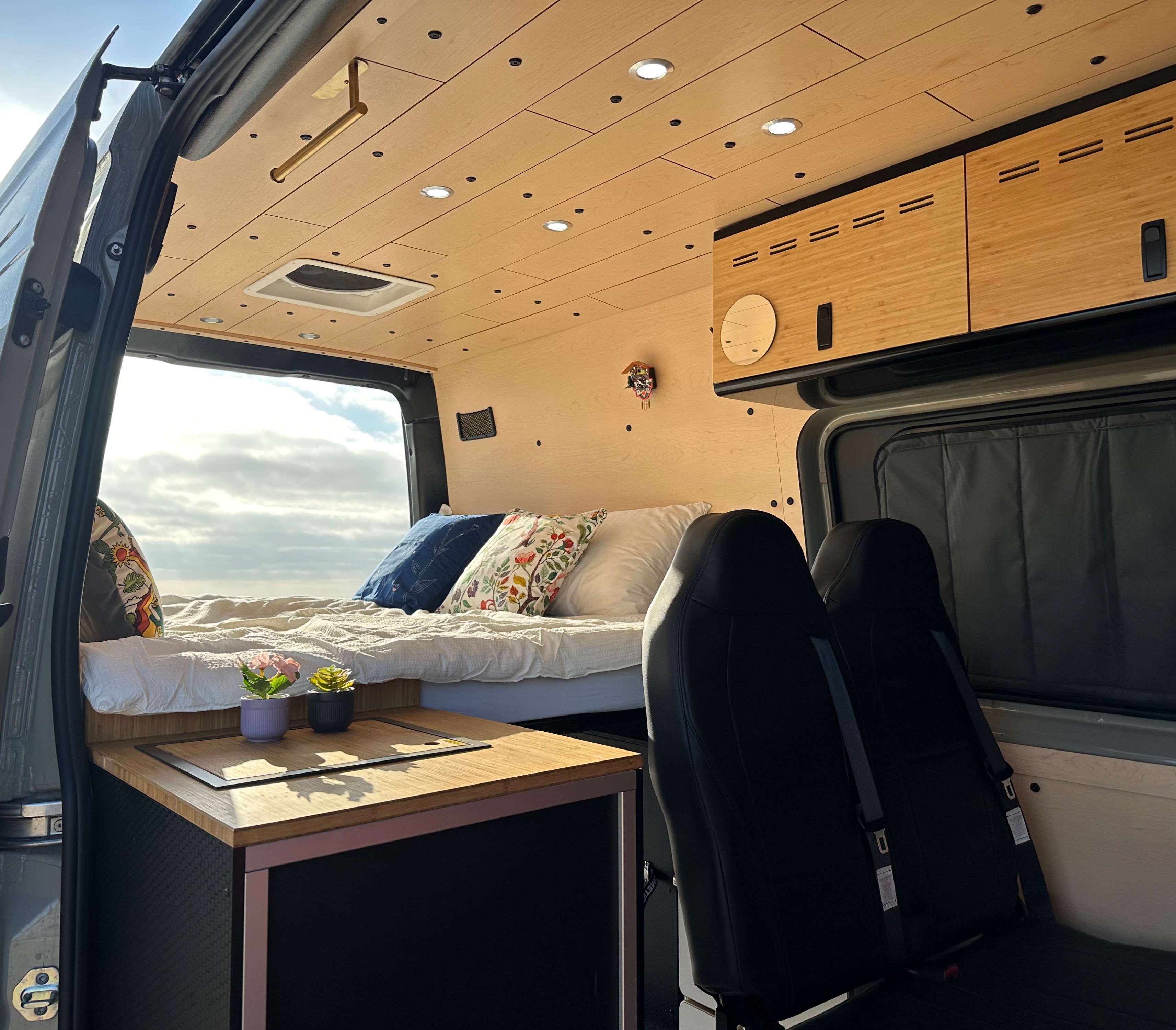 Inviting RV rental interior: wood-paneled ceiling, comfy bed, small kitchen, and front seats with a sky view.. Mercedes 144 2024