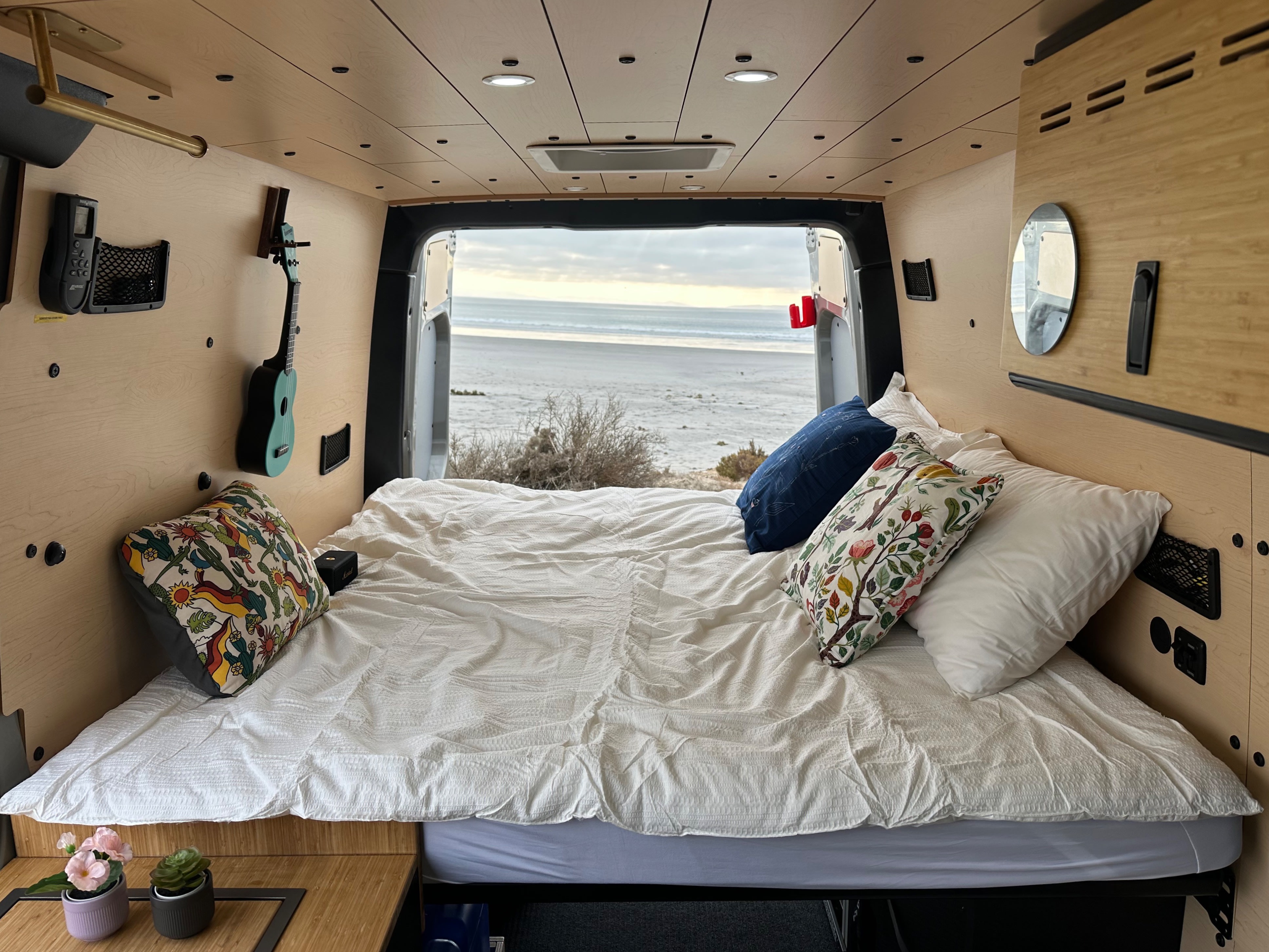 Inside a comfy RV rental, featuring a bed overlooking a calm beach. Perfect for relaxing travel & van life.. Mercedes 144 2024