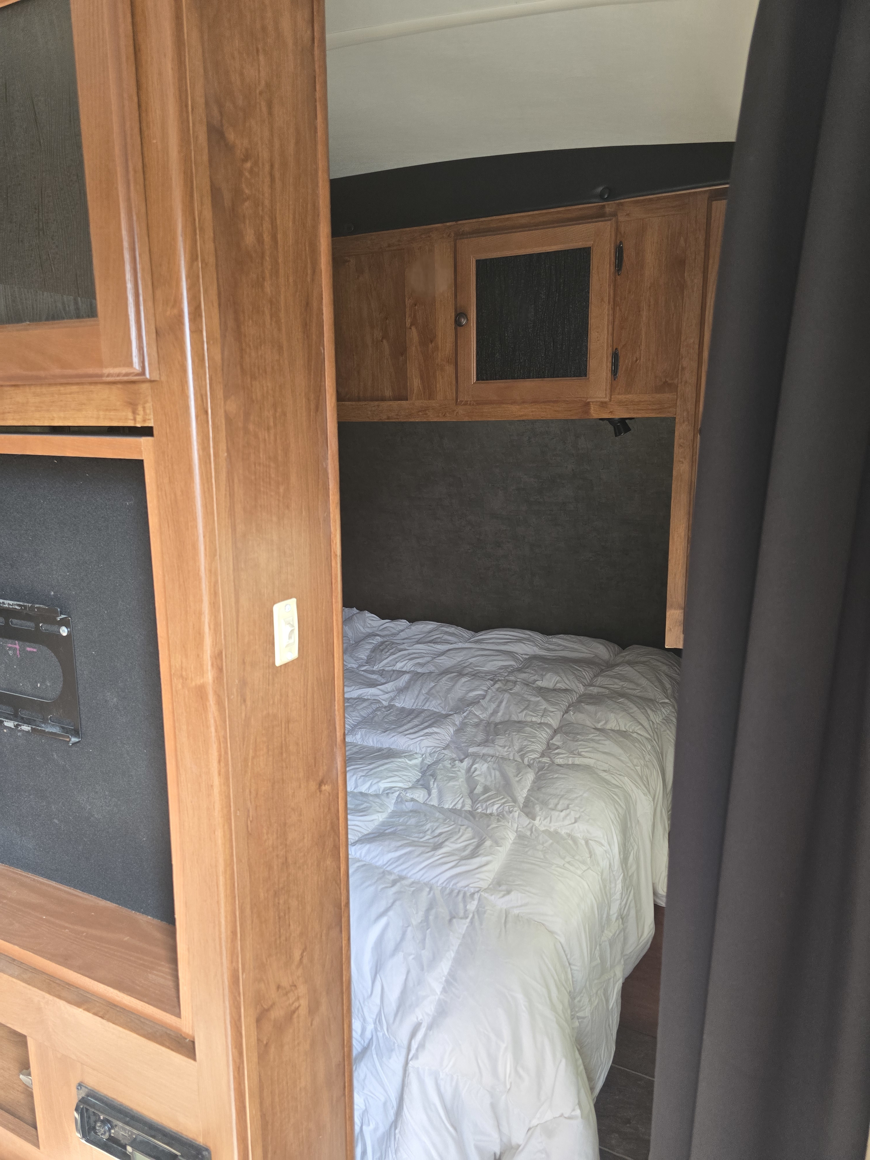 Inside an RV rental, featuring a cozy bedroom with a white comforter, wood cabinets, and a dark privacy curtain.. Heartland Wilderness 2015