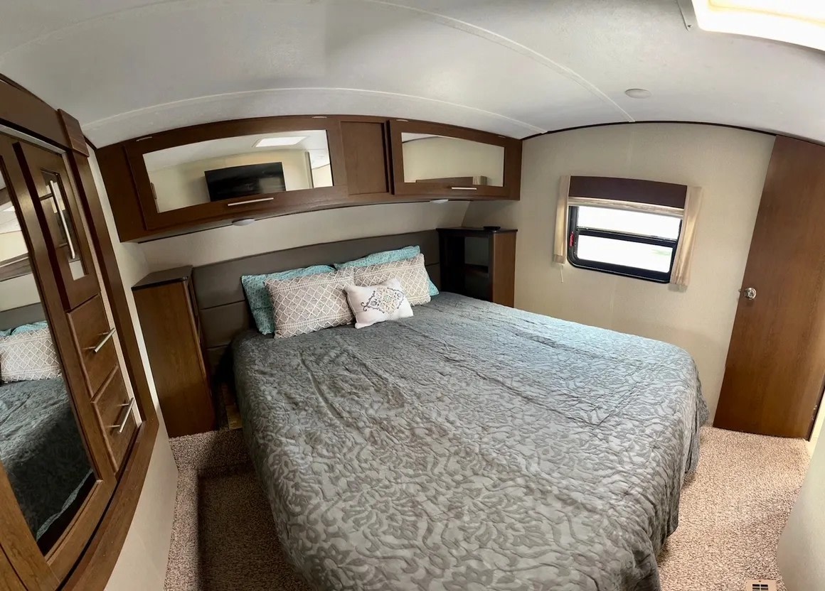 RV rental bedroom: a large bed with a grey patterned comforter, overhead mirrored cabinets, bedside tables, window, and built-in wardrobe.. Forest River Impression 2018