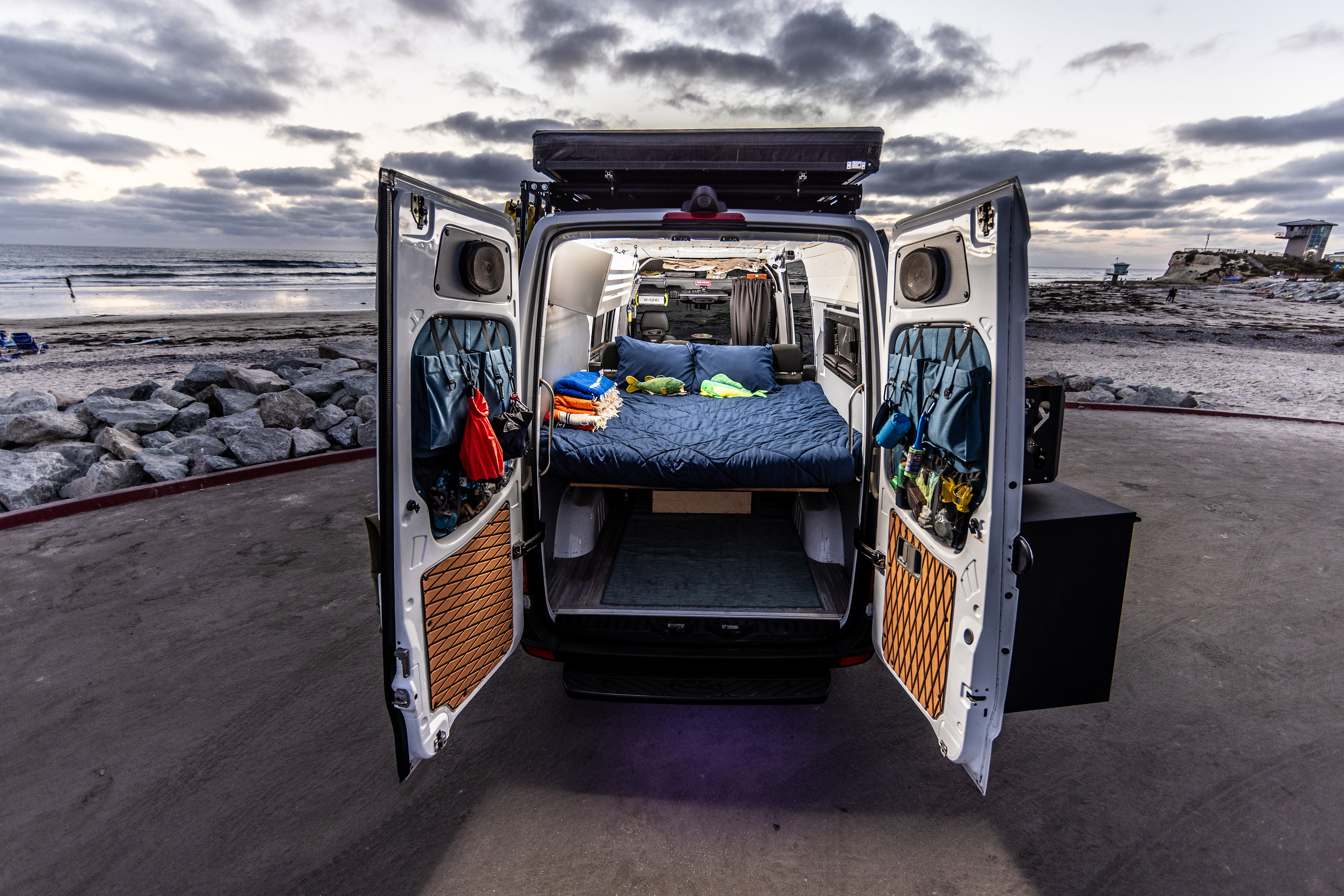 This RV rental camper van, parked beachside, reveals a cozy bed and organized interior through its open rear doors.. Mercedes-Benz Sprinter 2018