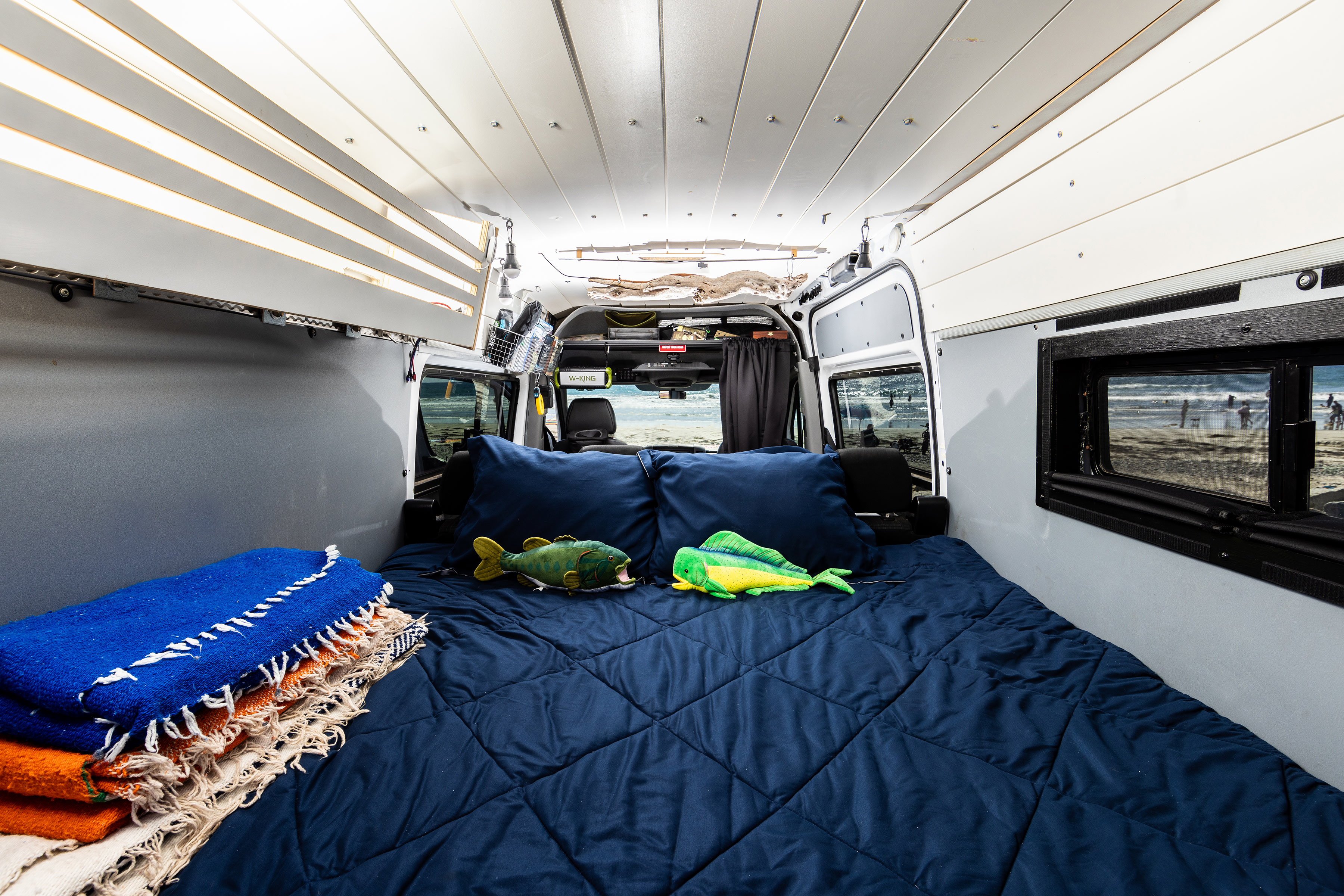 This cozy rv rental interior features a comfy bed with fish toys, stacked blankets, and open doors revealing a beach view.. Mercedes-Benz Sprinter 2018