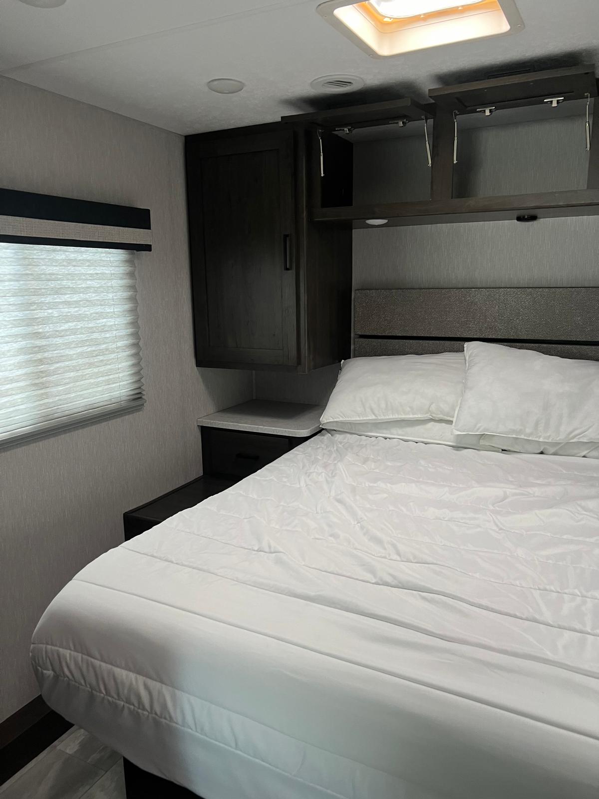 RV rental bedroom with white bedding, dark wood cabinets, and a window with blinds, offering a comfortable space.. Forest River Coachmen Pursuit 2023