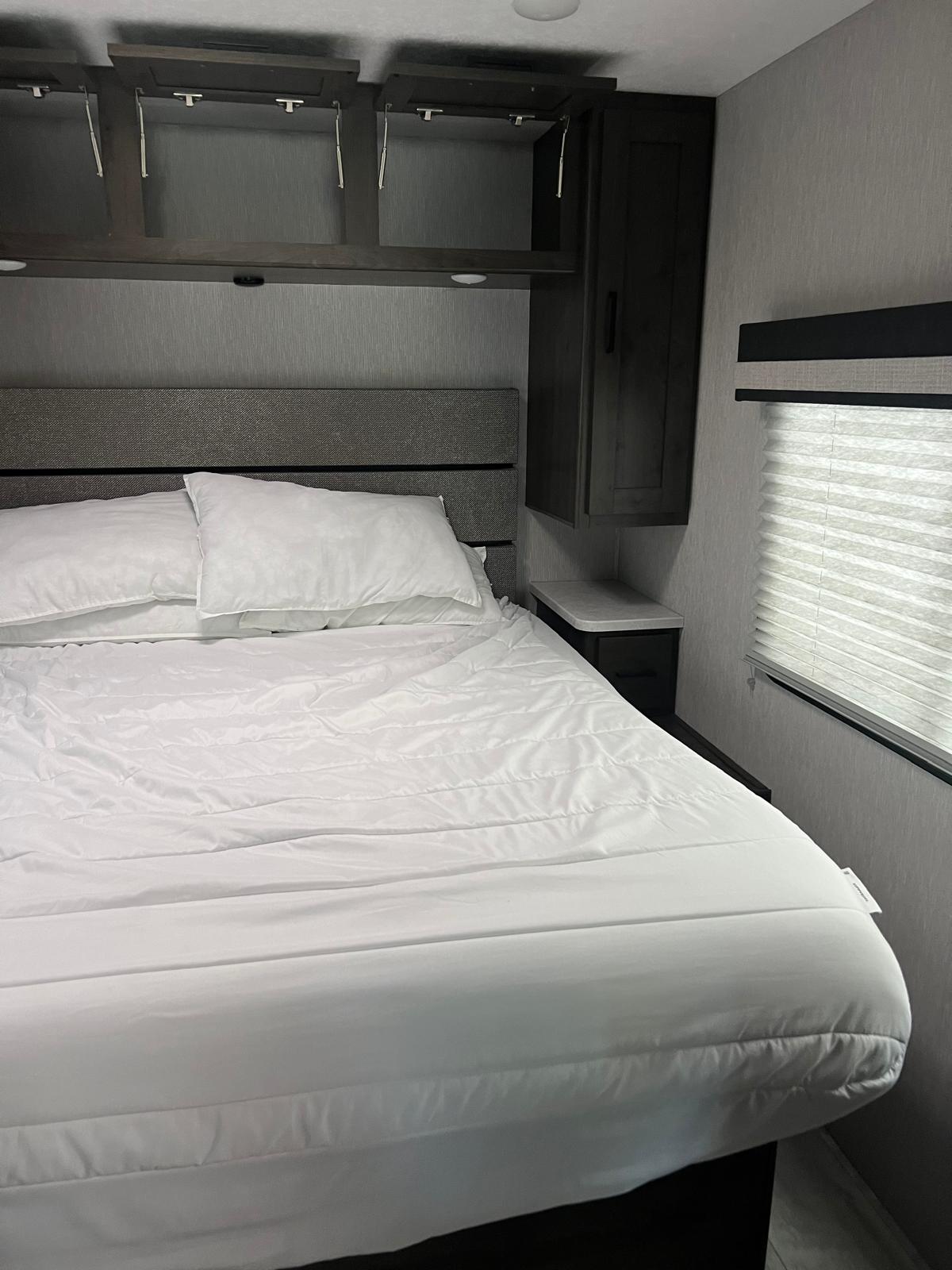 Cozy RV rental bedroom featuring a comfortable bed with white linens, overhead cabinets, and a window with blinds.. Forest River Coachmen Pursuit 2023